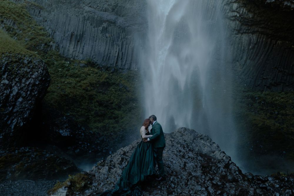 Silhouettes embrace in front of a towering waterfall during a dramatic photoshoot.