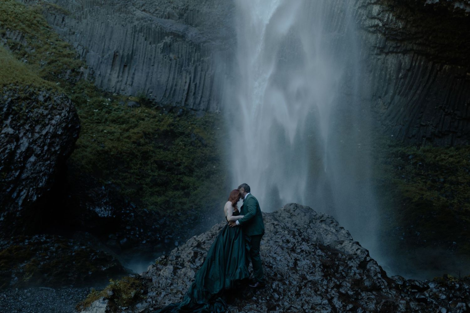 Silhouettes embrace in front of a towering waterfall during a dramatic photoshoot.