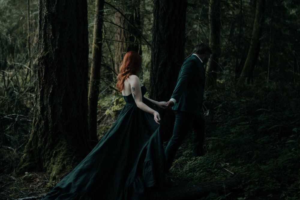 People walk along a forest path surrounded by tall pine trees in moody lighting.