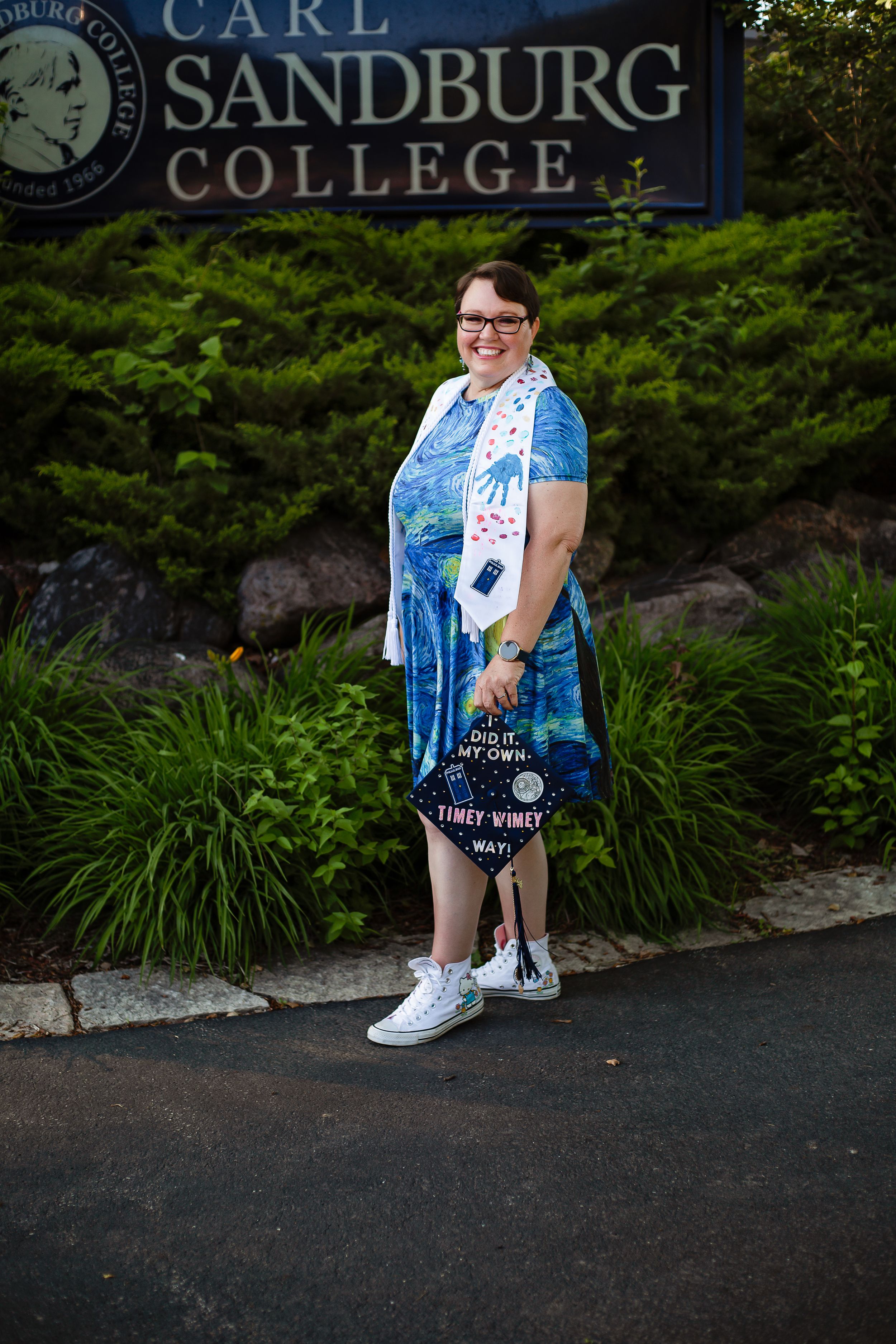 Graduate posing by Carl Sandburg College sign wearing blue graduation attire.