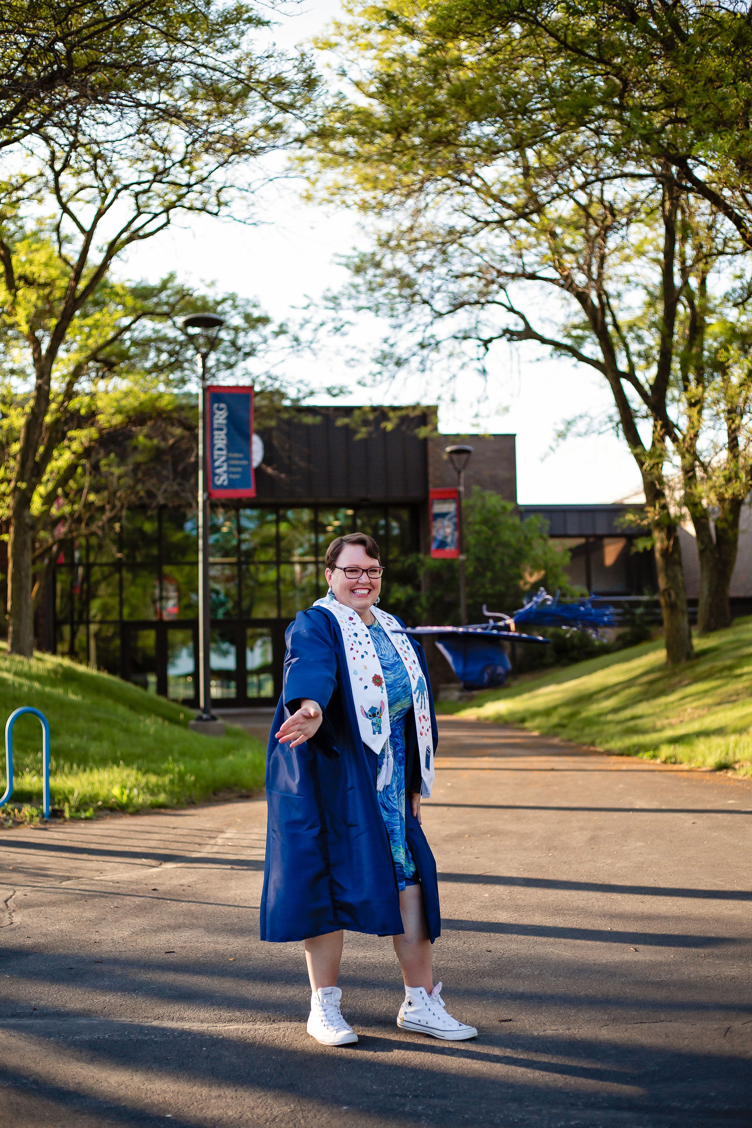 Graduate in blue cap and gown poses on campus pathway surrounded by trees.