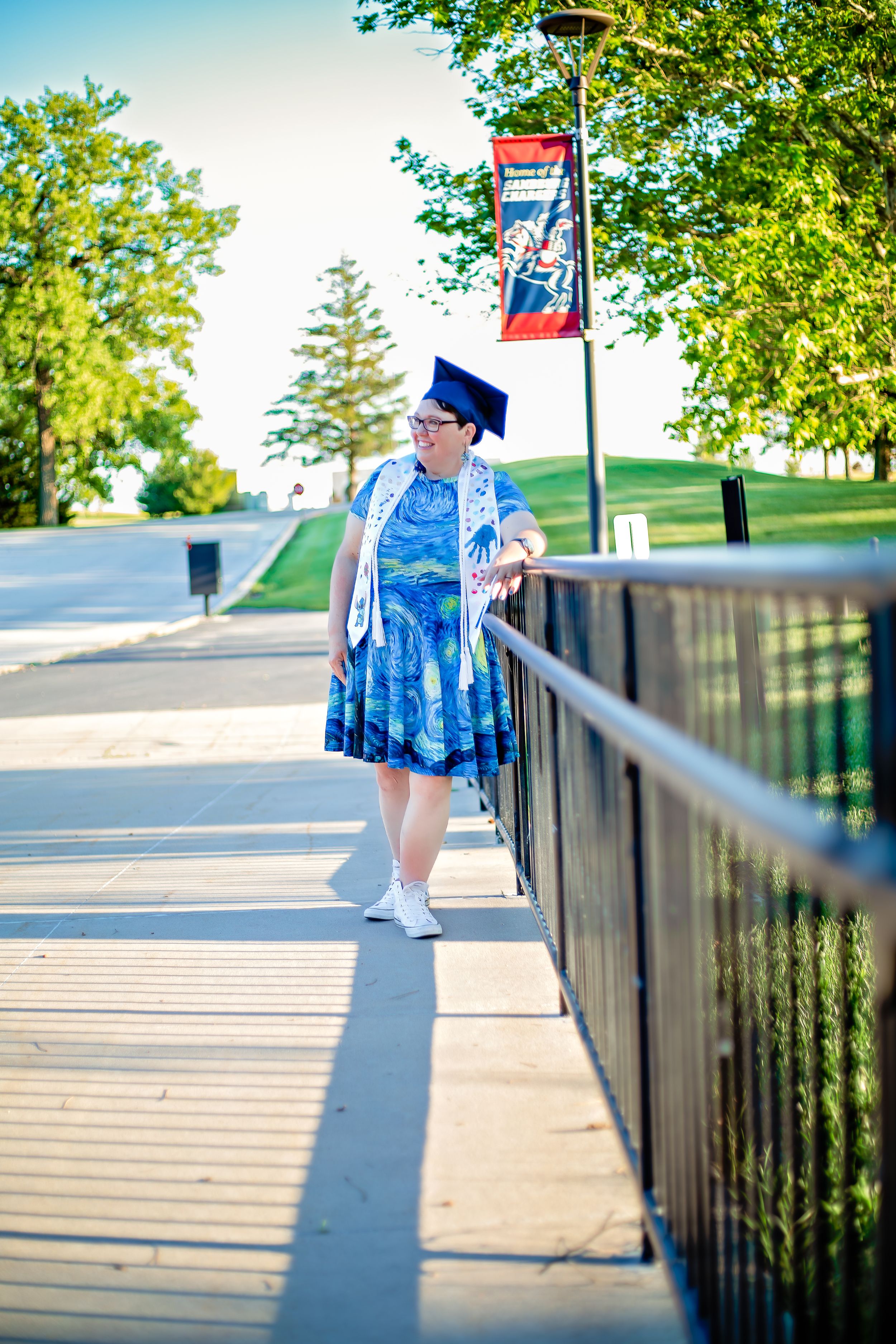 Graduate in blue dress and cap standing on walkway with decorative banner at college campus.