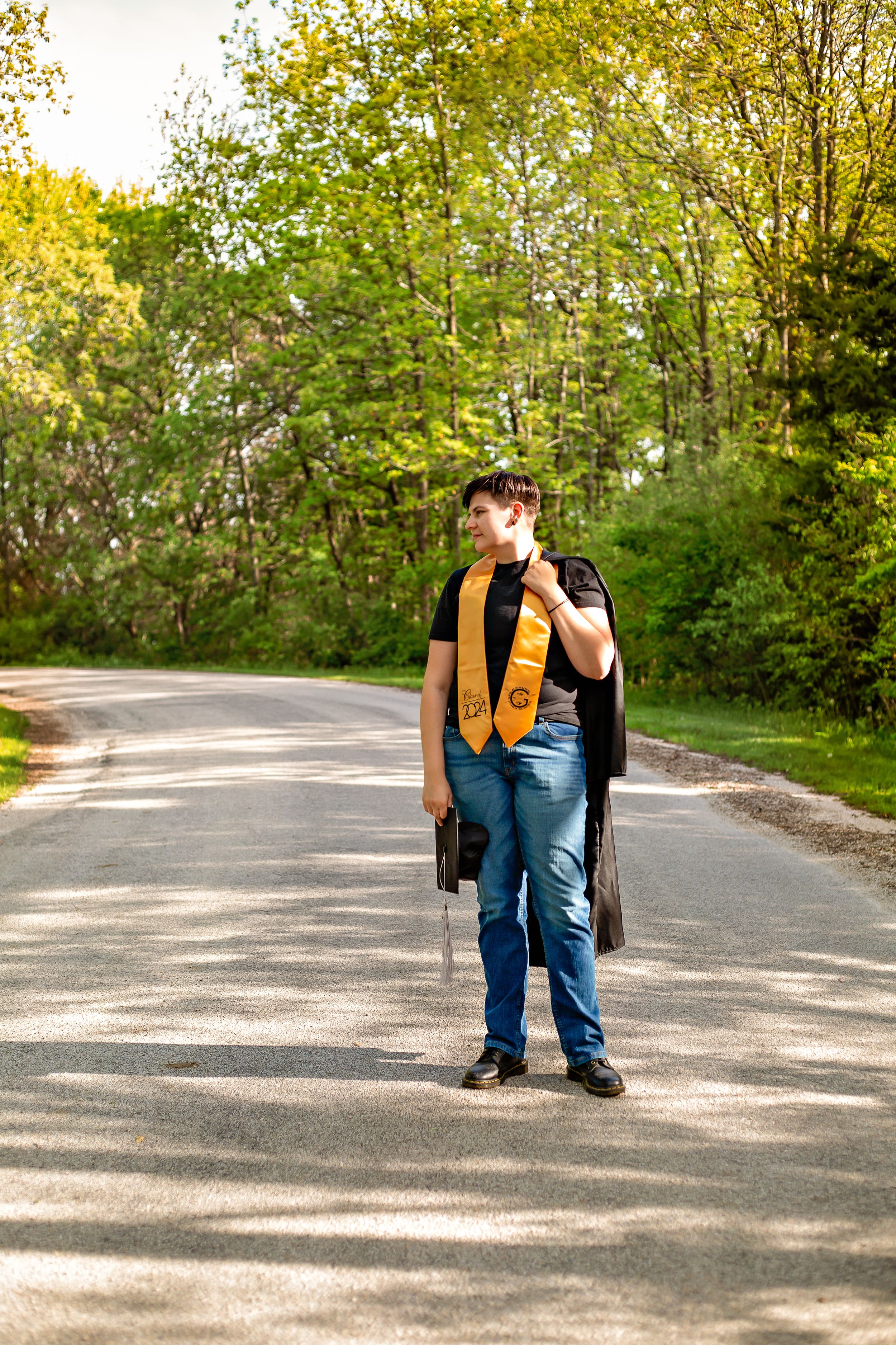 Graduate wearing yellow stole and carrying graduation gown walks on tree-lined path.