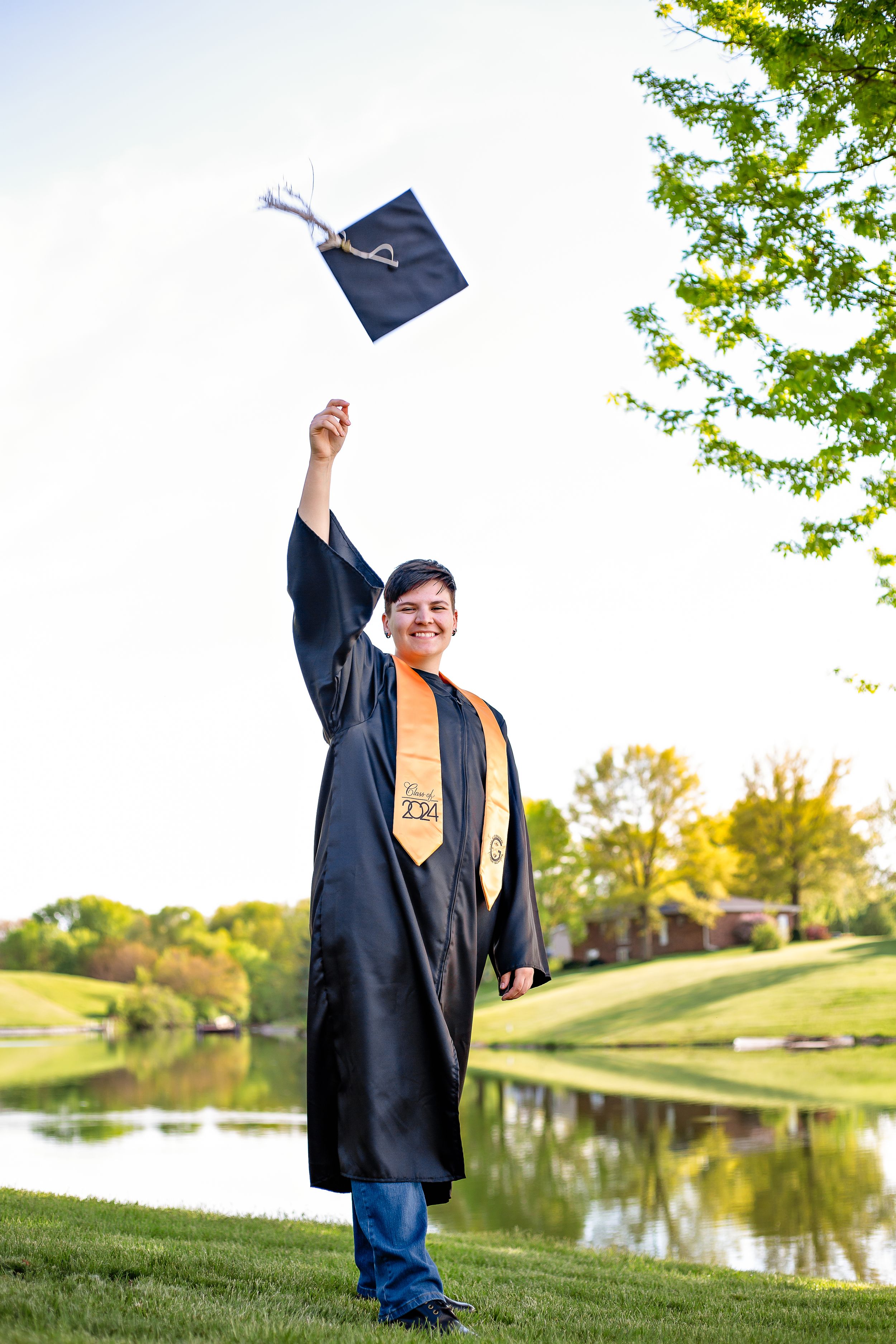 Graduate throwing mortarboard cap in air by lake during graduation celebration.
