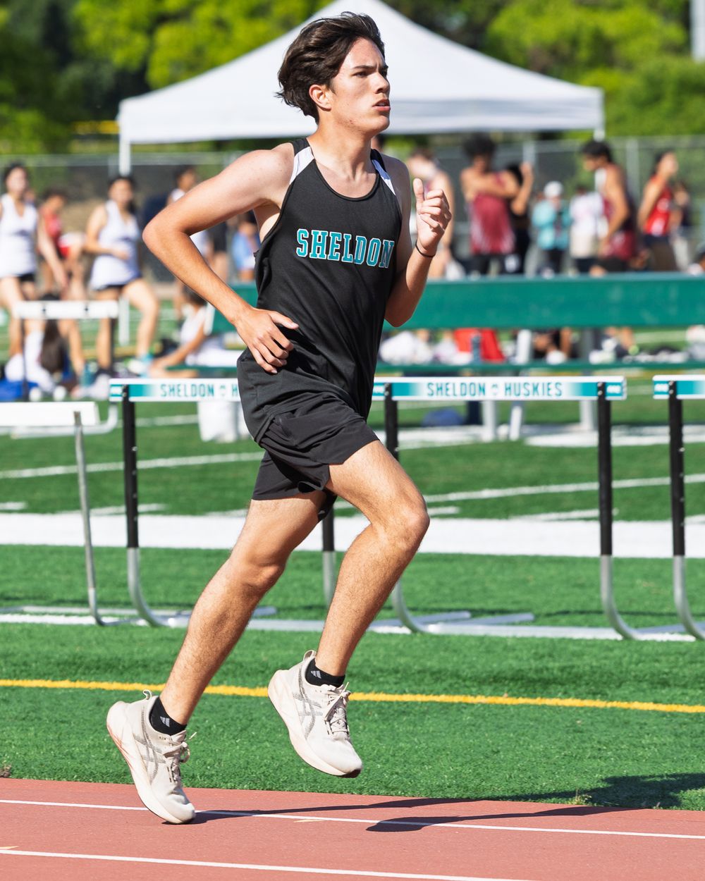 Sheldon Huskies track athlete runs on a red track during a hurdles competition on a sunny day.