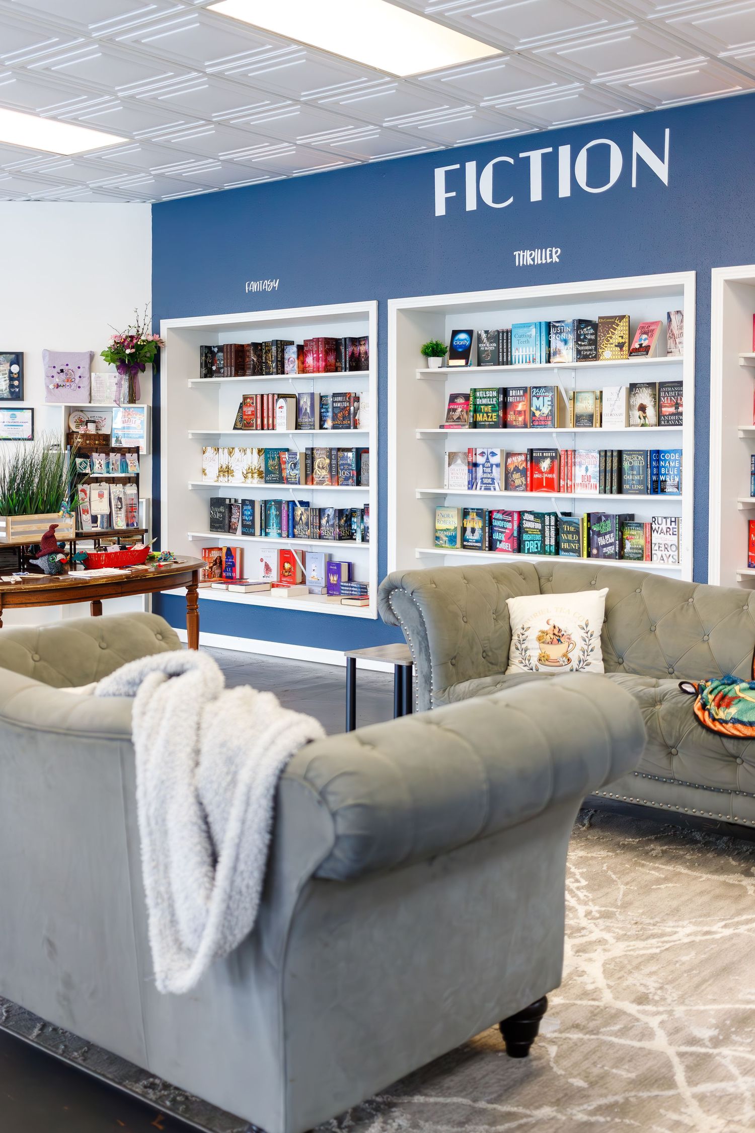 Modern bookstore fiction section with gray armchairs and shelves against a blue accent wall.