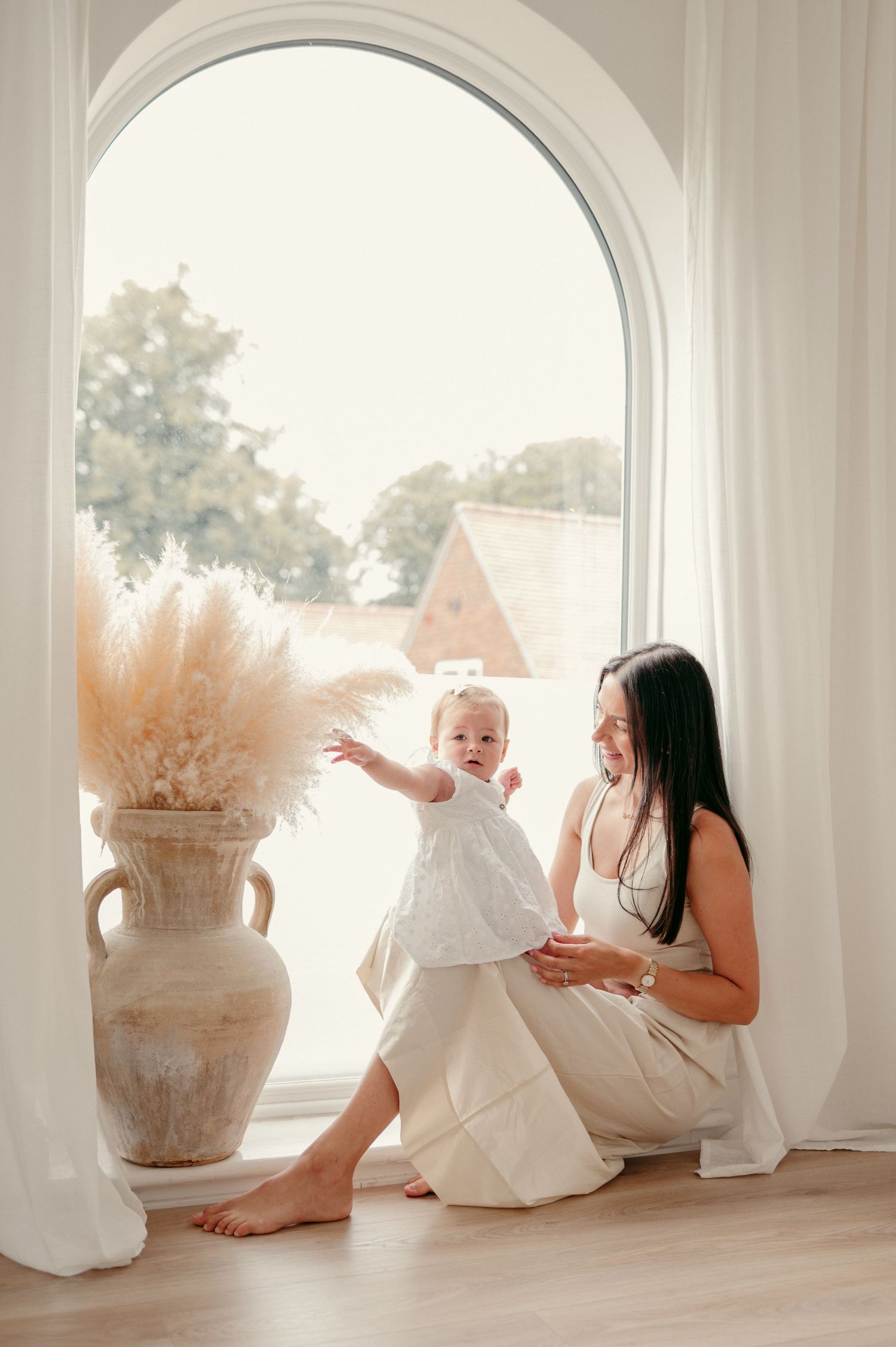A bright indoor scene with parent and baby in white outfits near an arched window and decorative vase.
