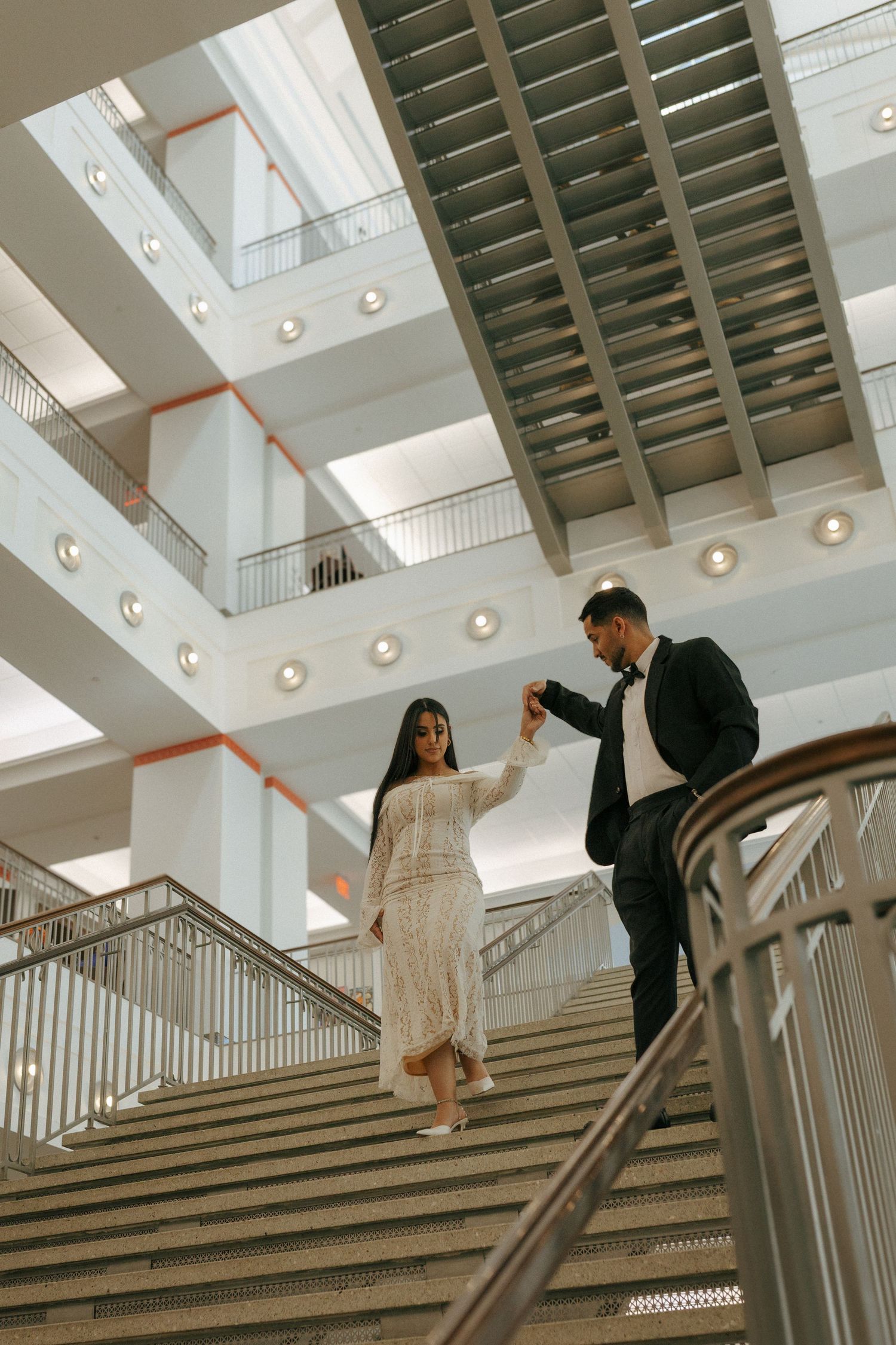 A couple descends a grand staircase in an architectural building with natural light streaming through skylights above.