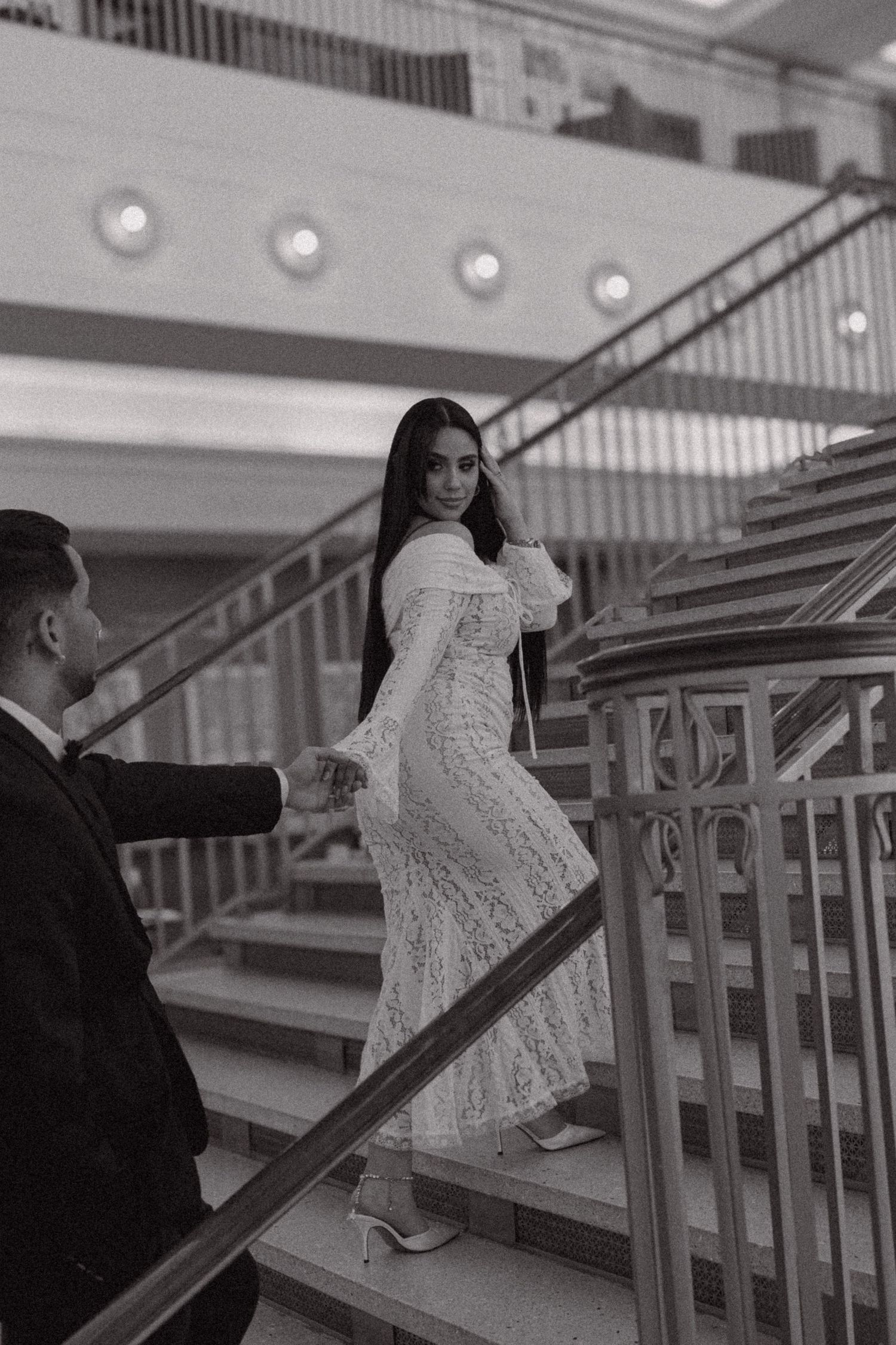 A couple in formal attire ascends an elegant staircase in a black and white photo, with her wearing a lace dress.