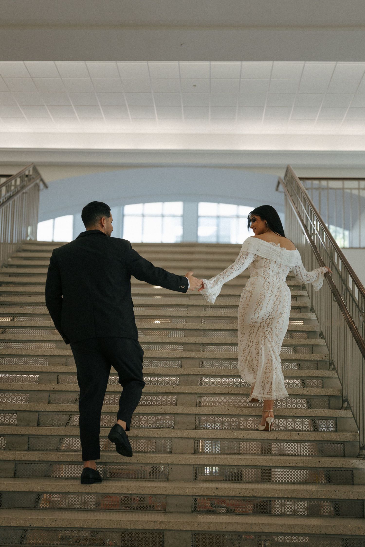 A couple in wedding attire holds hands while ascending a grand staircase, shot from behind in an elegant indoor setting.