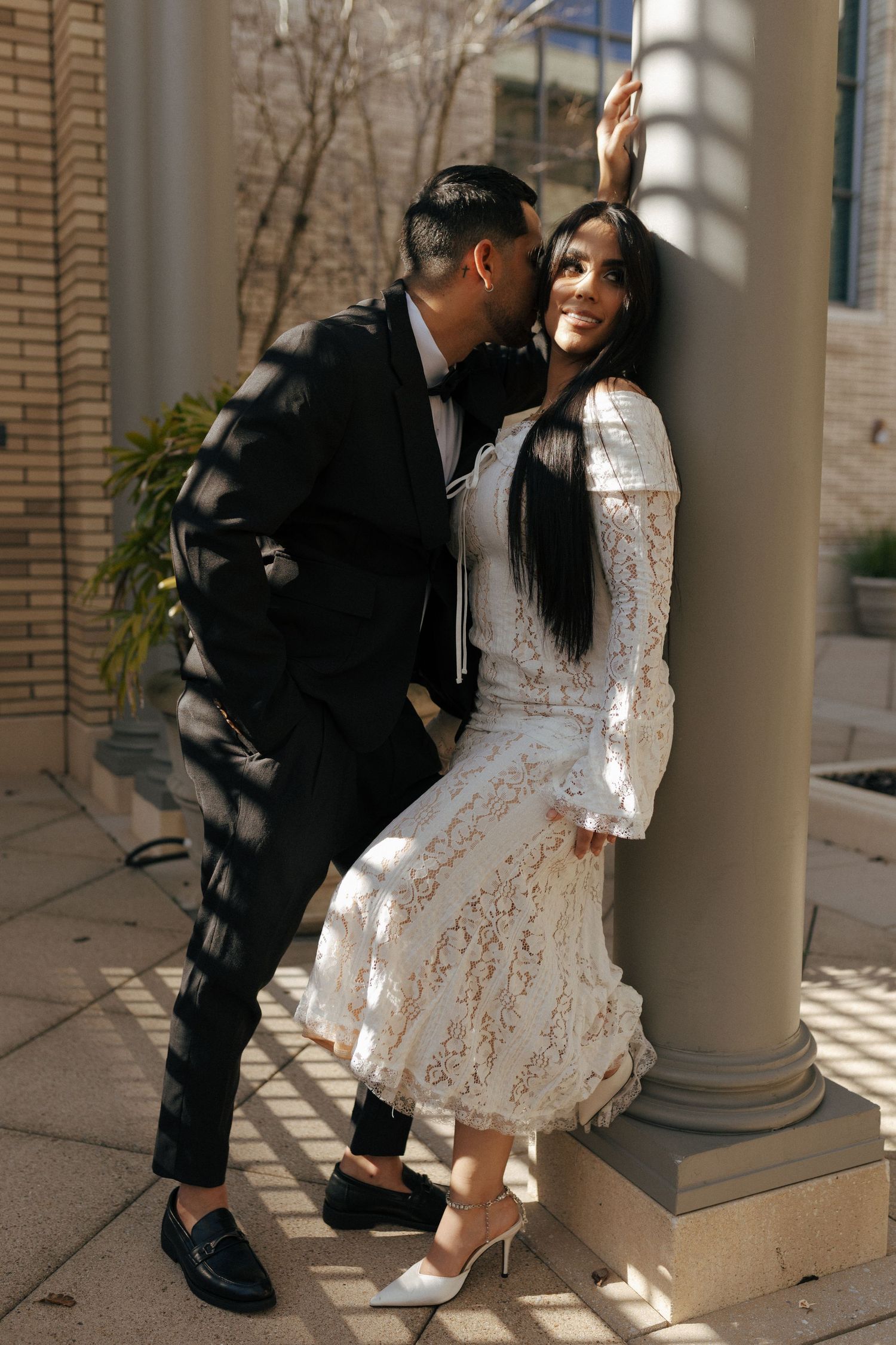 A couple dressed in black and white formal attire poses playfully against a column in warm sunlight.