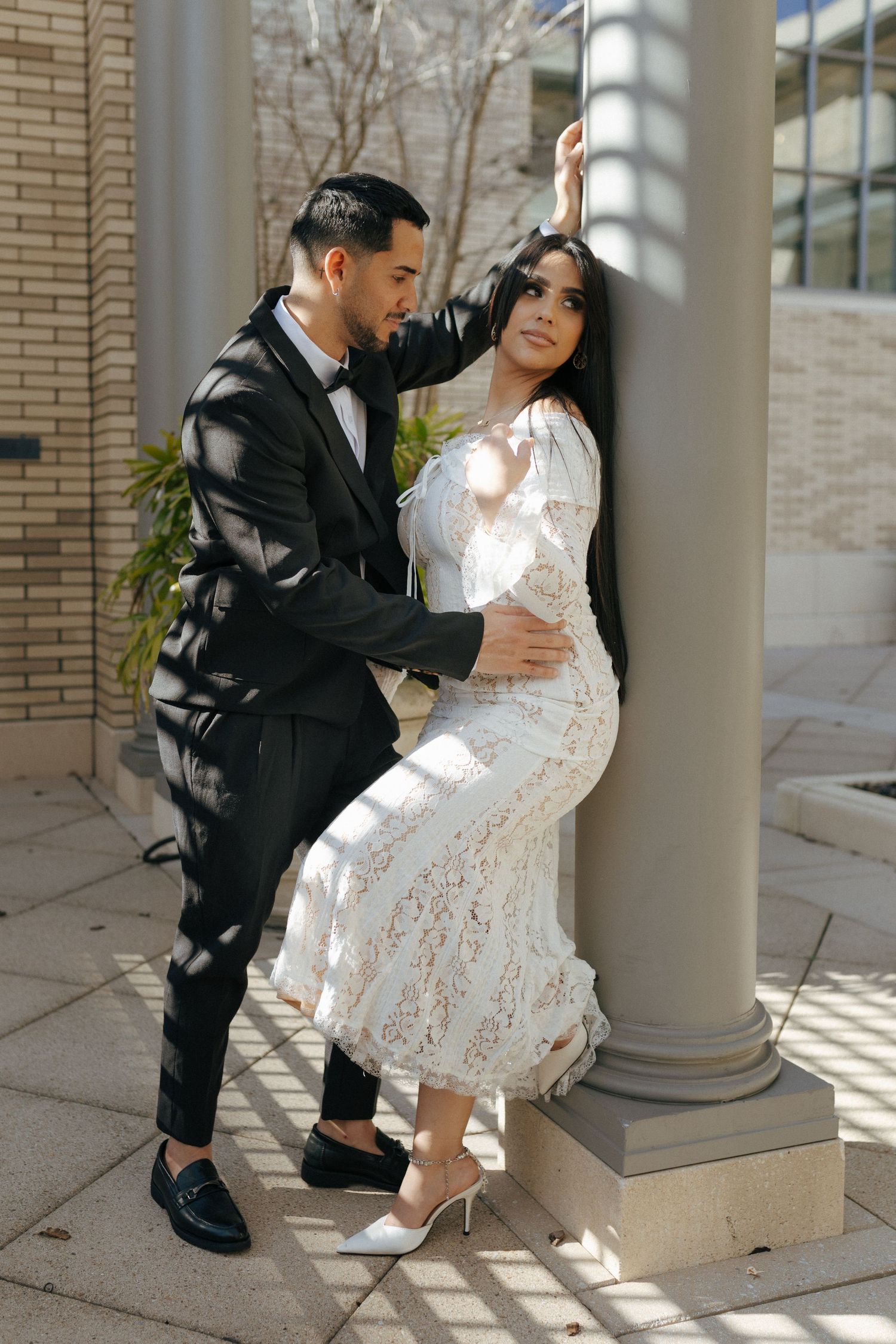 Couple in formal attire poses romantically by architectural column with sunlight casting dramatic shadows.