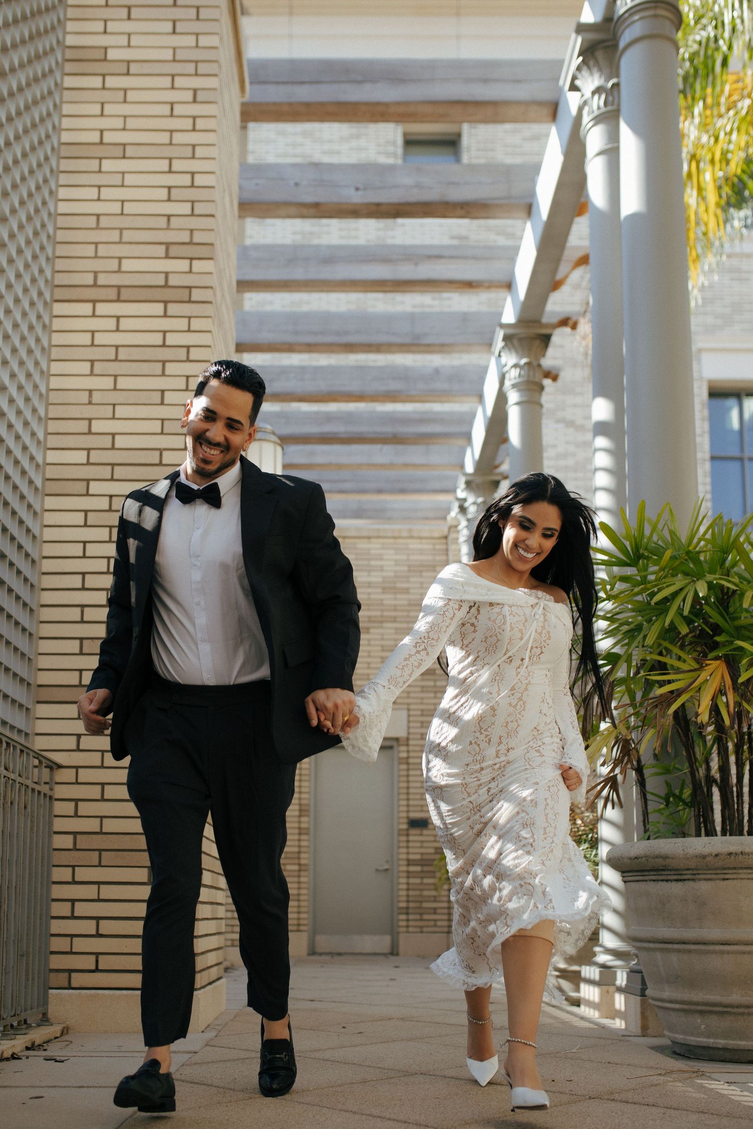 Elegantly dressed couple in formal attire hold hands while walking together along a covered outdoor walkway.