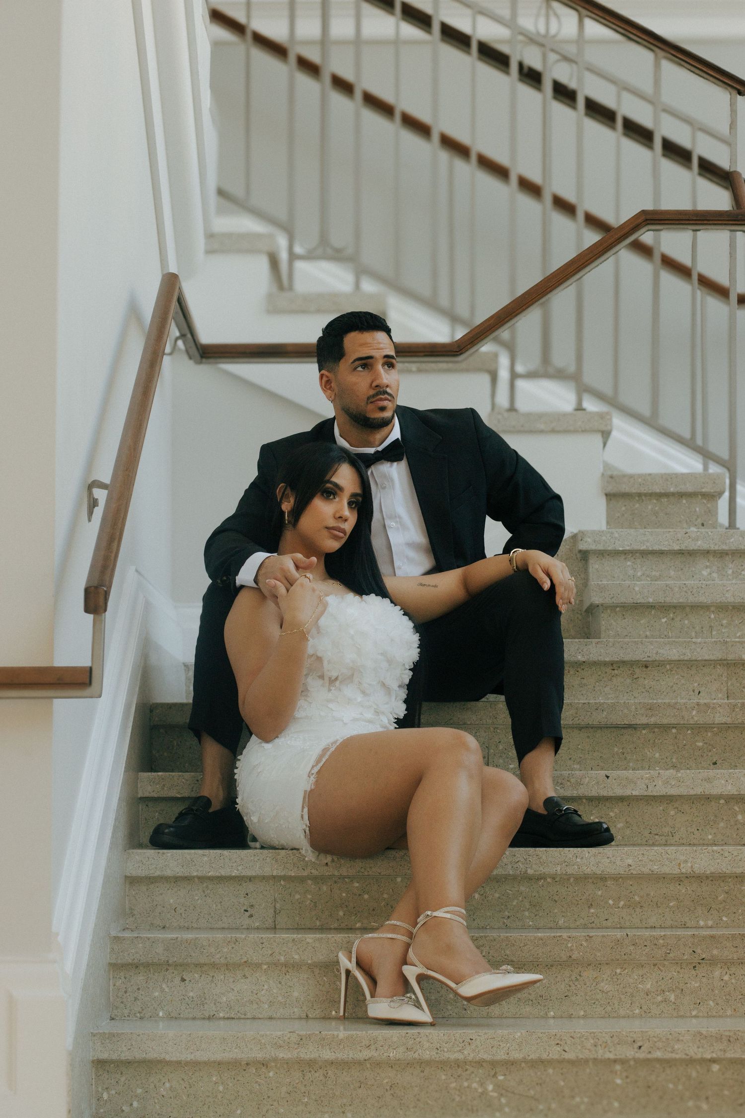 A couple in formal attire poses intimately on elegant white stairs, with her in a short white dress and him in a black suit.