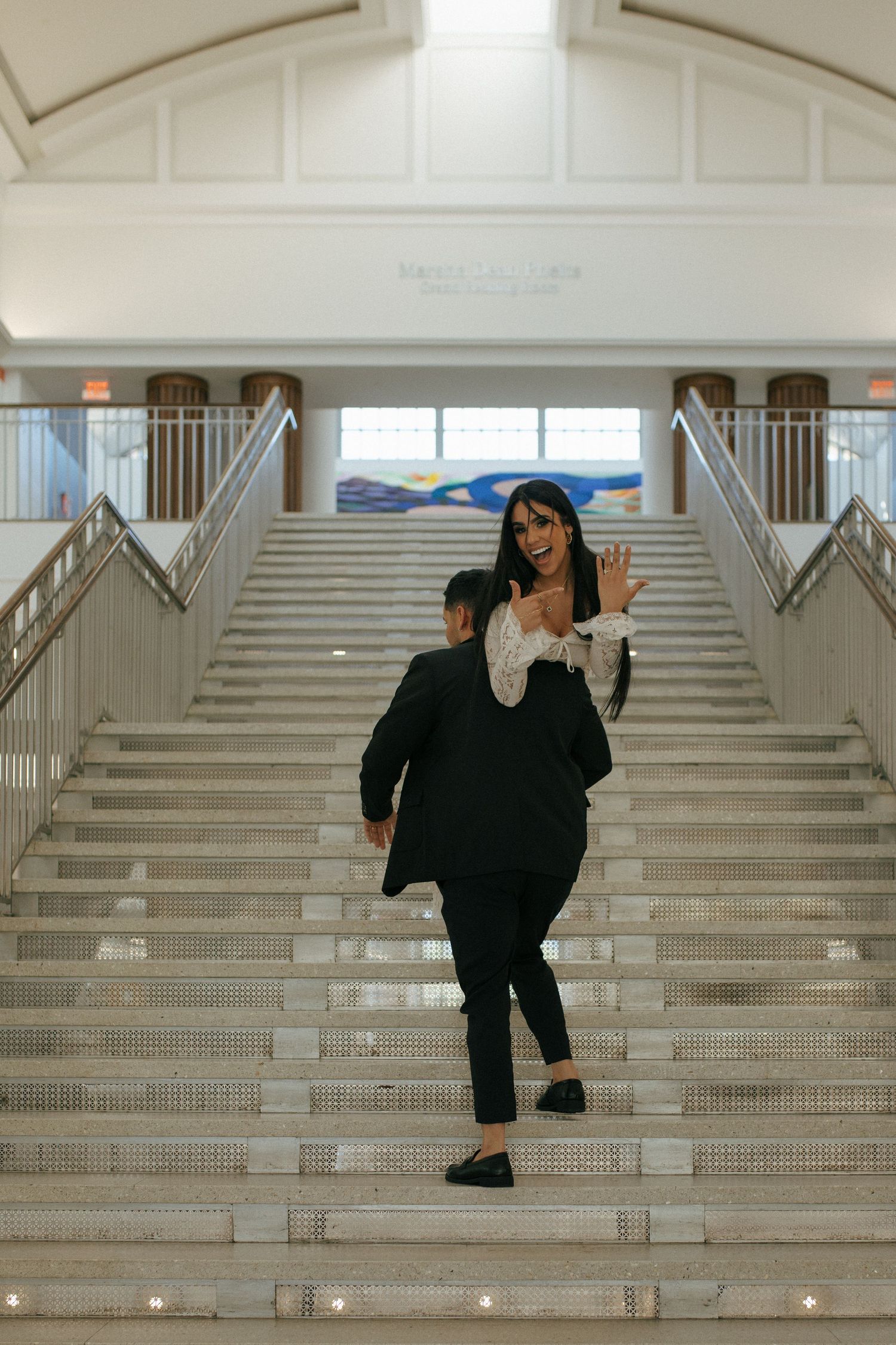 A couple descends a grand white staircase in a building with arched ceilings and natural lighting.