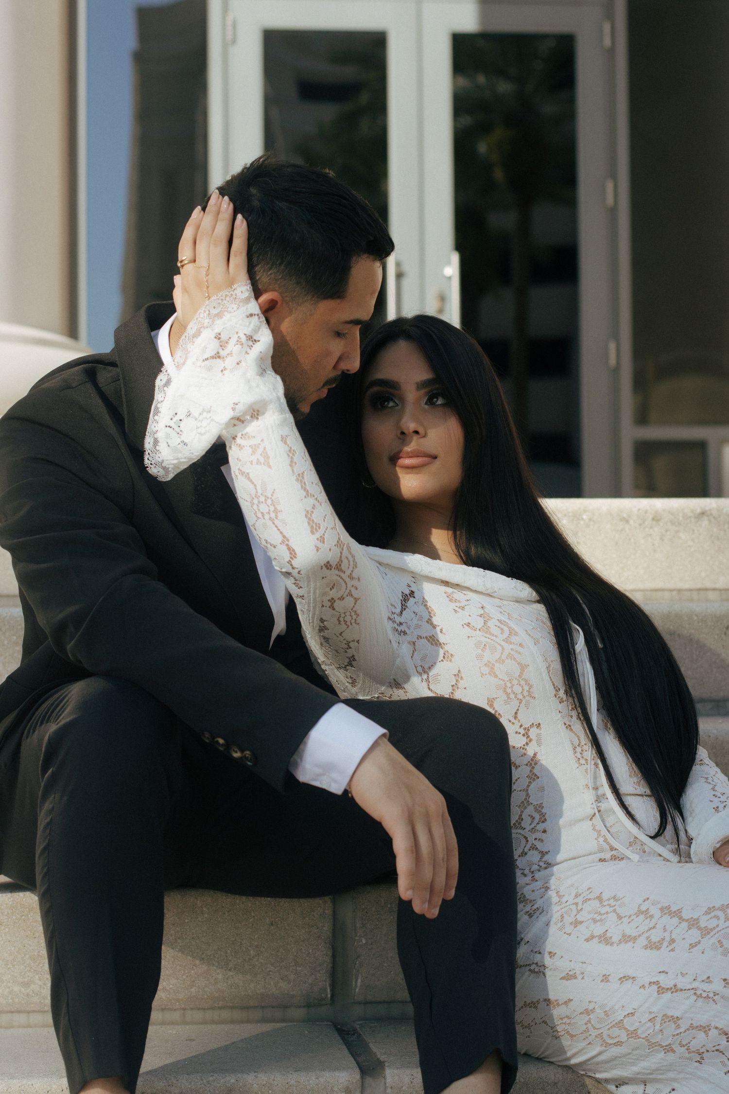 Romantic couple in elegant attire sharing an intimate moment on outdoor steps, with woman in white lace dress.