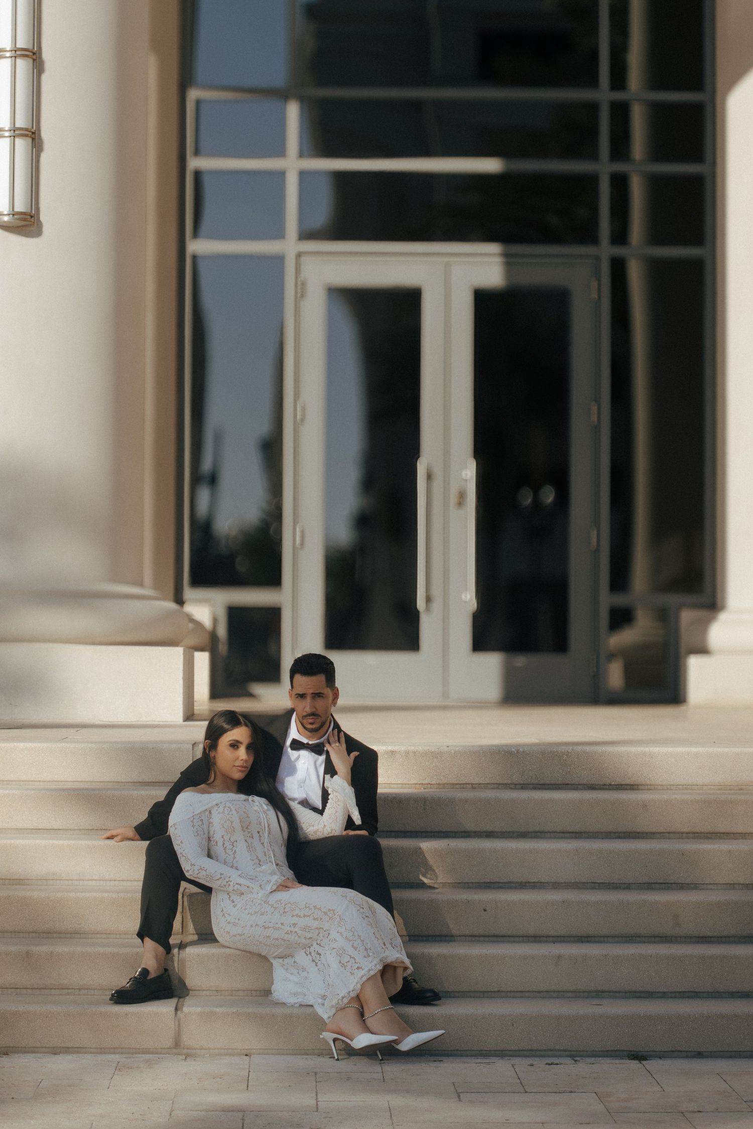A couple in formal wedding attire sits on elegant stone steps outside a building with glass doors.