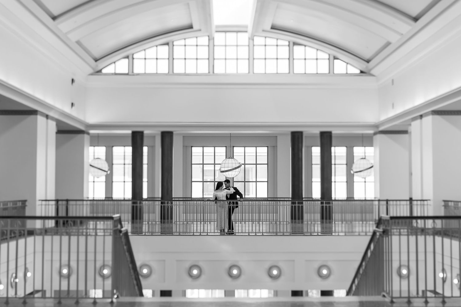 Black and white photograph of two silhouetted figures embracing on a balcony in an elegant interior with arched windows and railings.