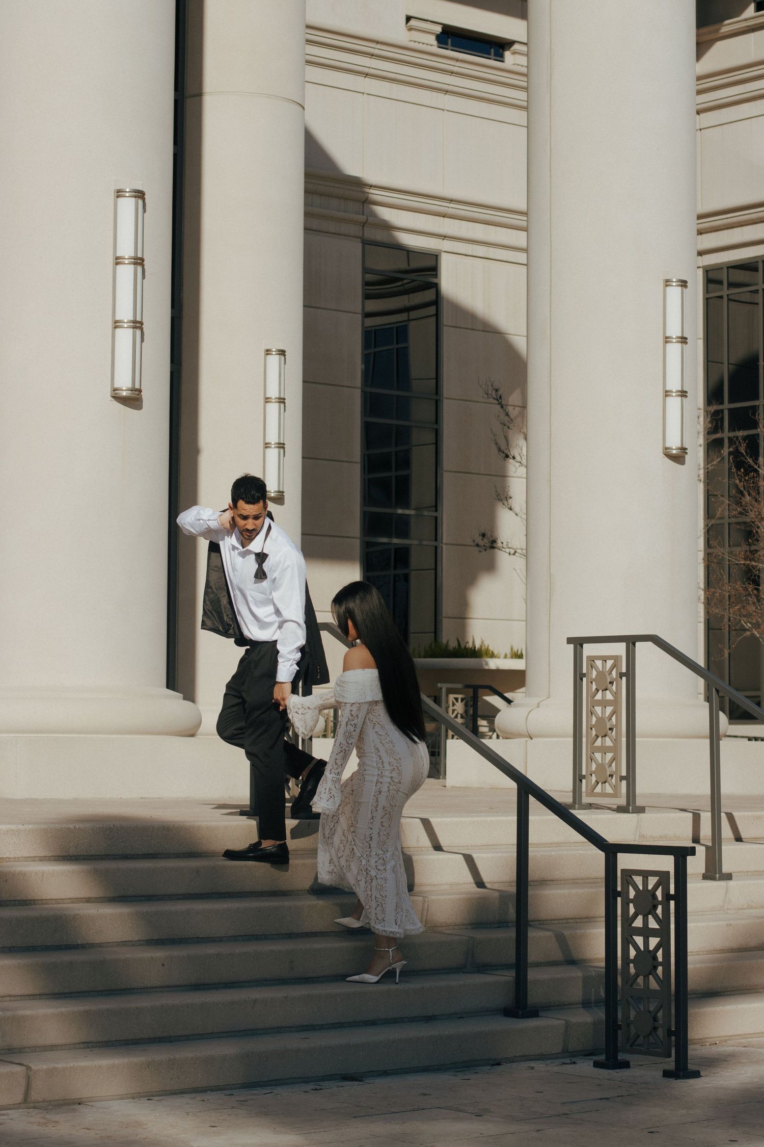 Couple interacting on elegant stone steps with white columns and decorative railings in warm sunlight.