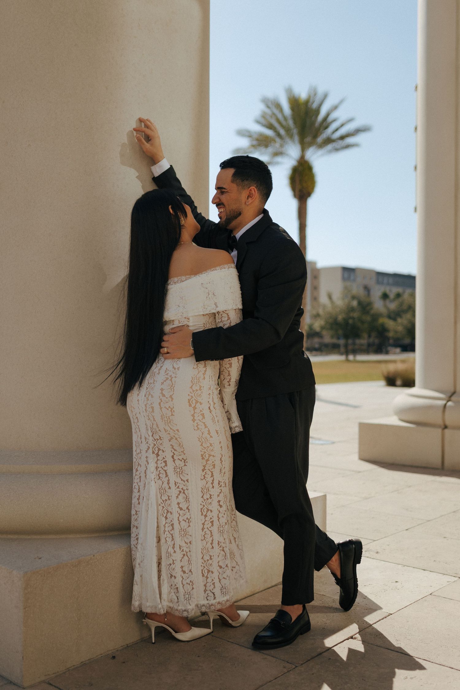 A couple embraces romantically on a sun-lit porch with a palm tree visible in the background.