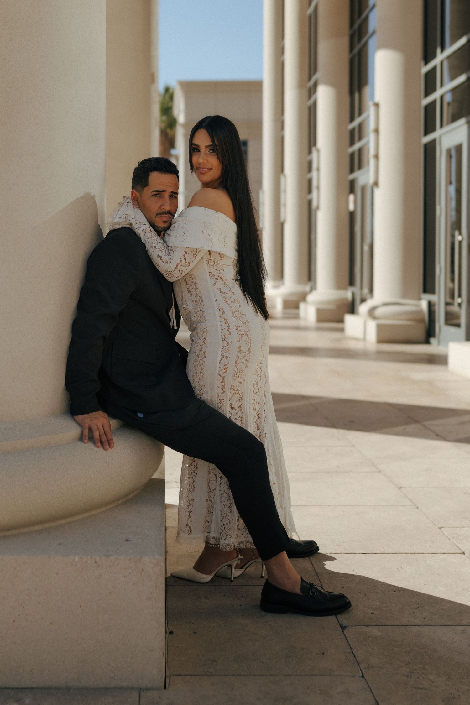 A romantic couple in formal attire poses against a classical building with columns in warm sunlight.