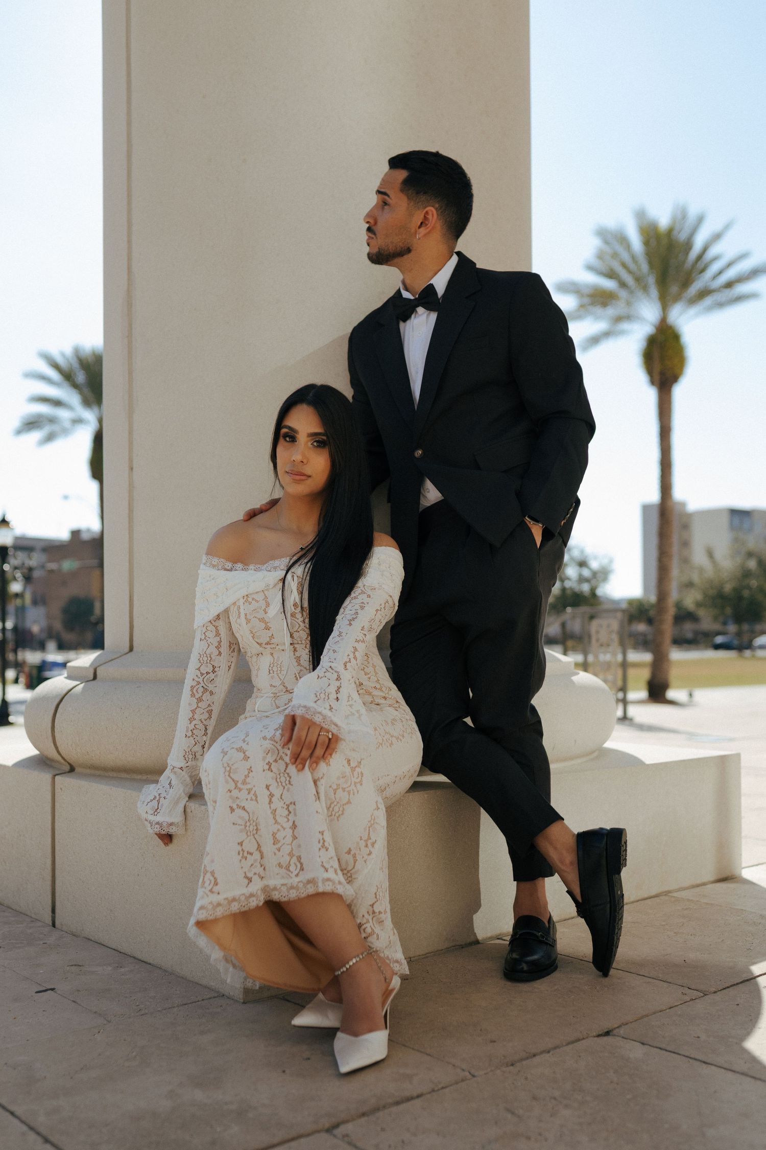 Elegant couple poses on marble steps in formal attire, white lace dress and black tuxedo against palm tree backdrop.