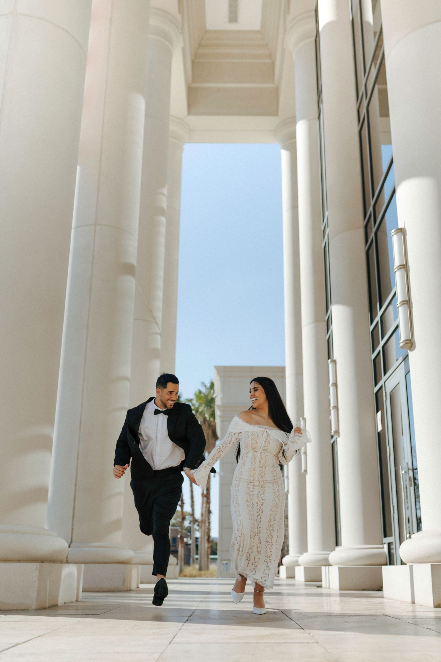 A couple in formal attire runs joyfully between elegant white columns at a grand venue.