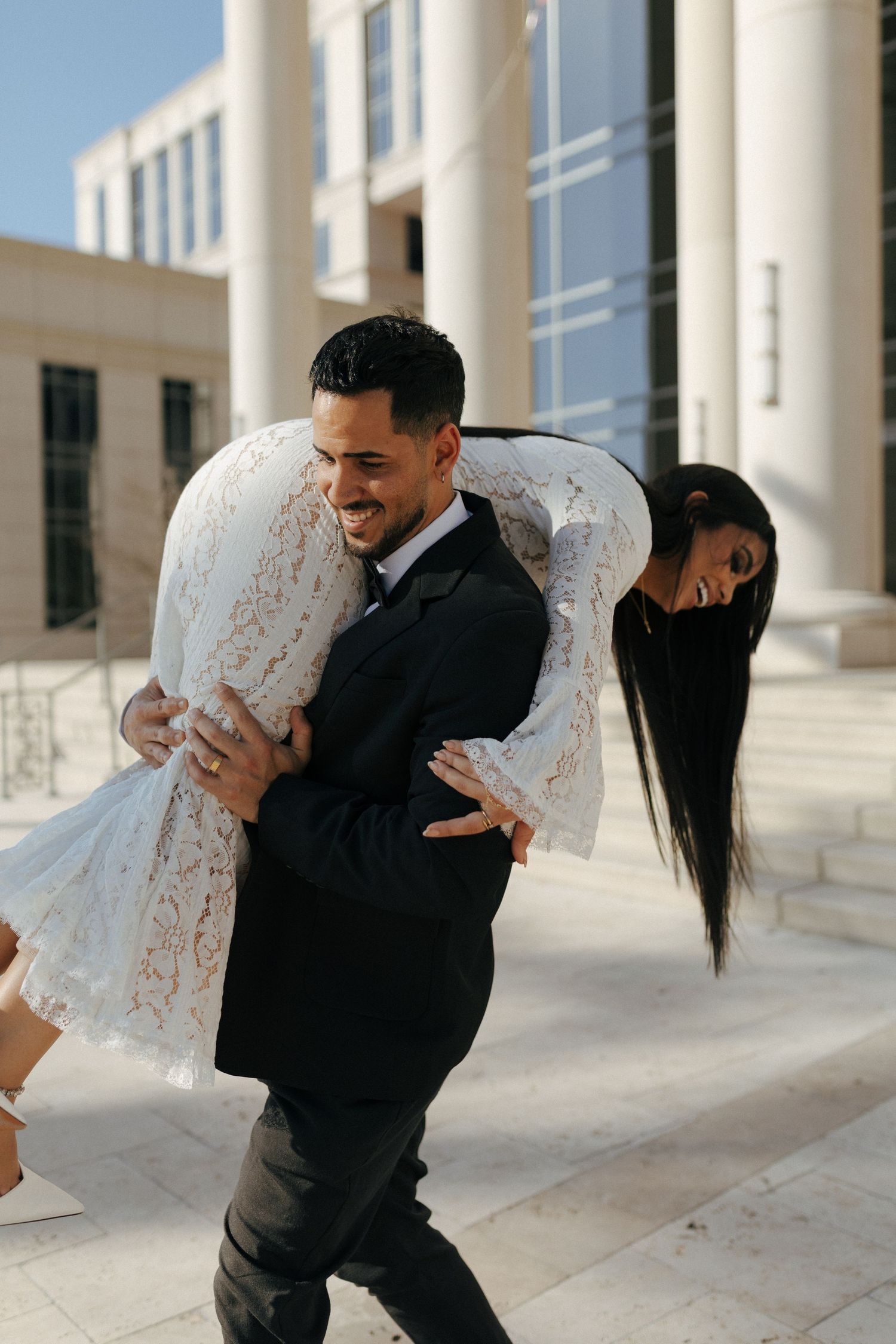 Joyful couple shares a playful moment on their wedding day outside a building with white columns.