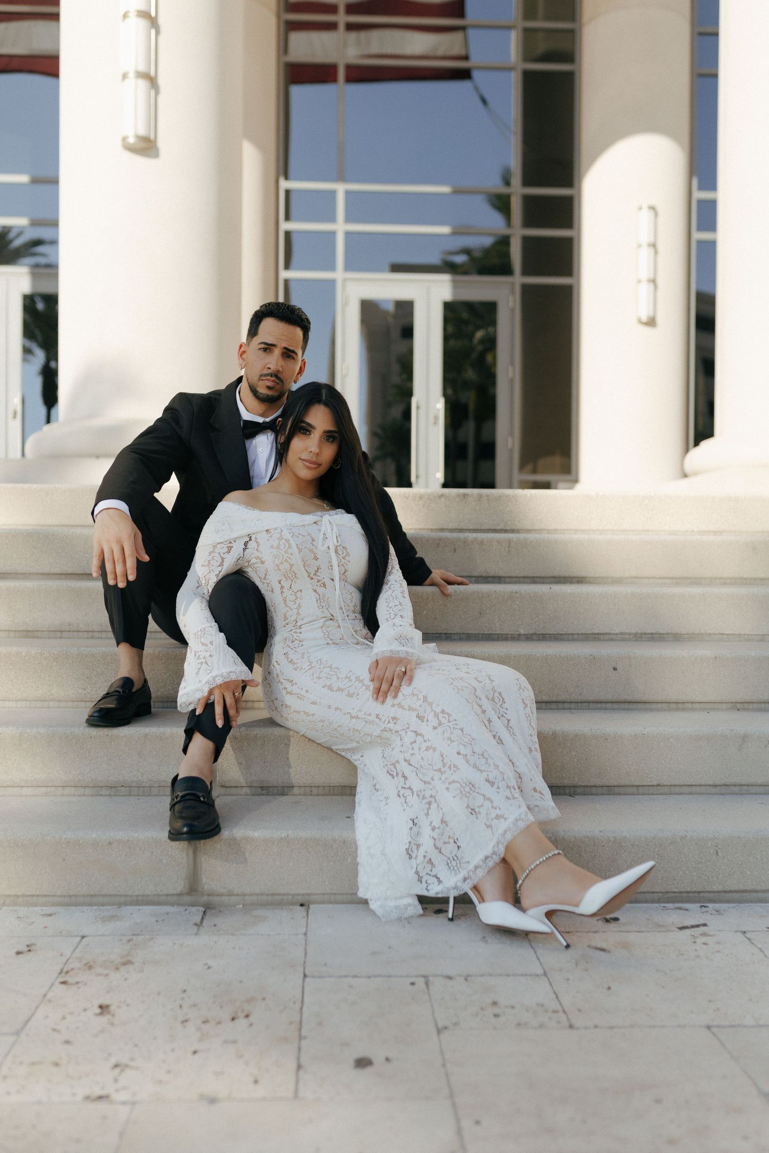 A couple poses on outdoor white marble steps, with the bride in a lace gown and the groom in black attire.