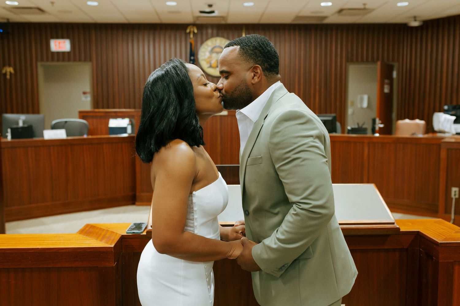 A couple shares a romantic kiss in a courthouse wearing formal white and grey attire during their wedding ceremony.