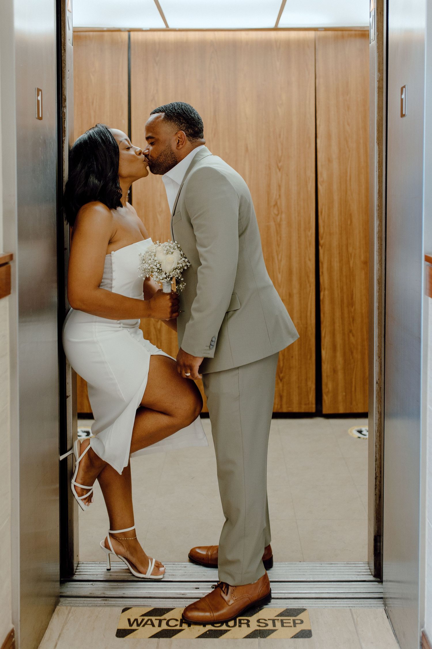 A romantic couple shares a kiss in an elevator while holding a small white bouquet at their wedding photoshoot.