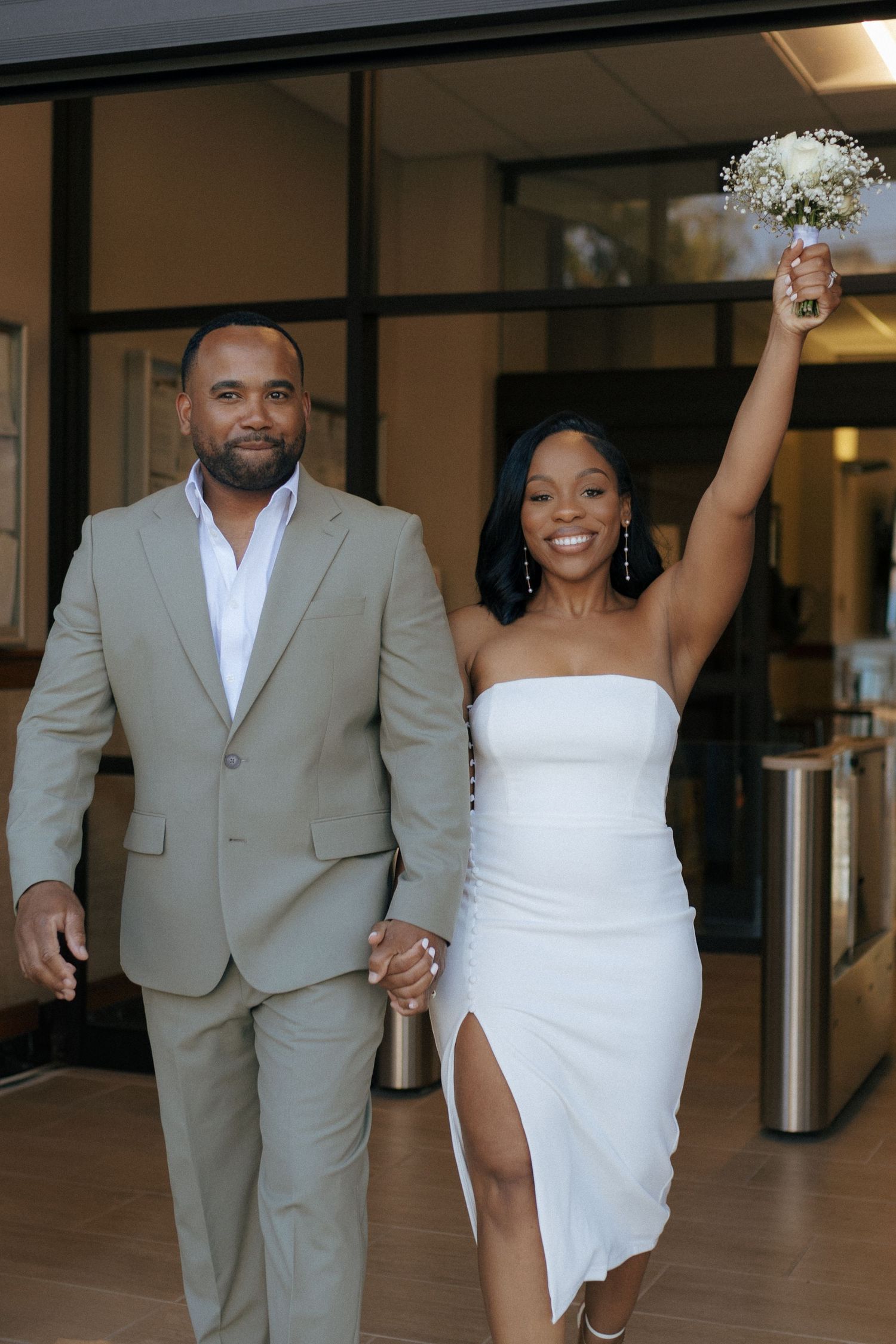 A celebratory couple walk together, with one wearing a tan suit and the other in a white dress holding a bouquet.