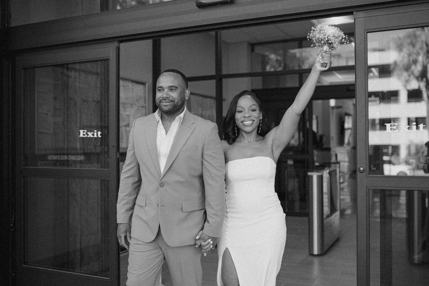 A joyful couple holds hands outside a building entrance, with the bride in white raising her hand in celebration.