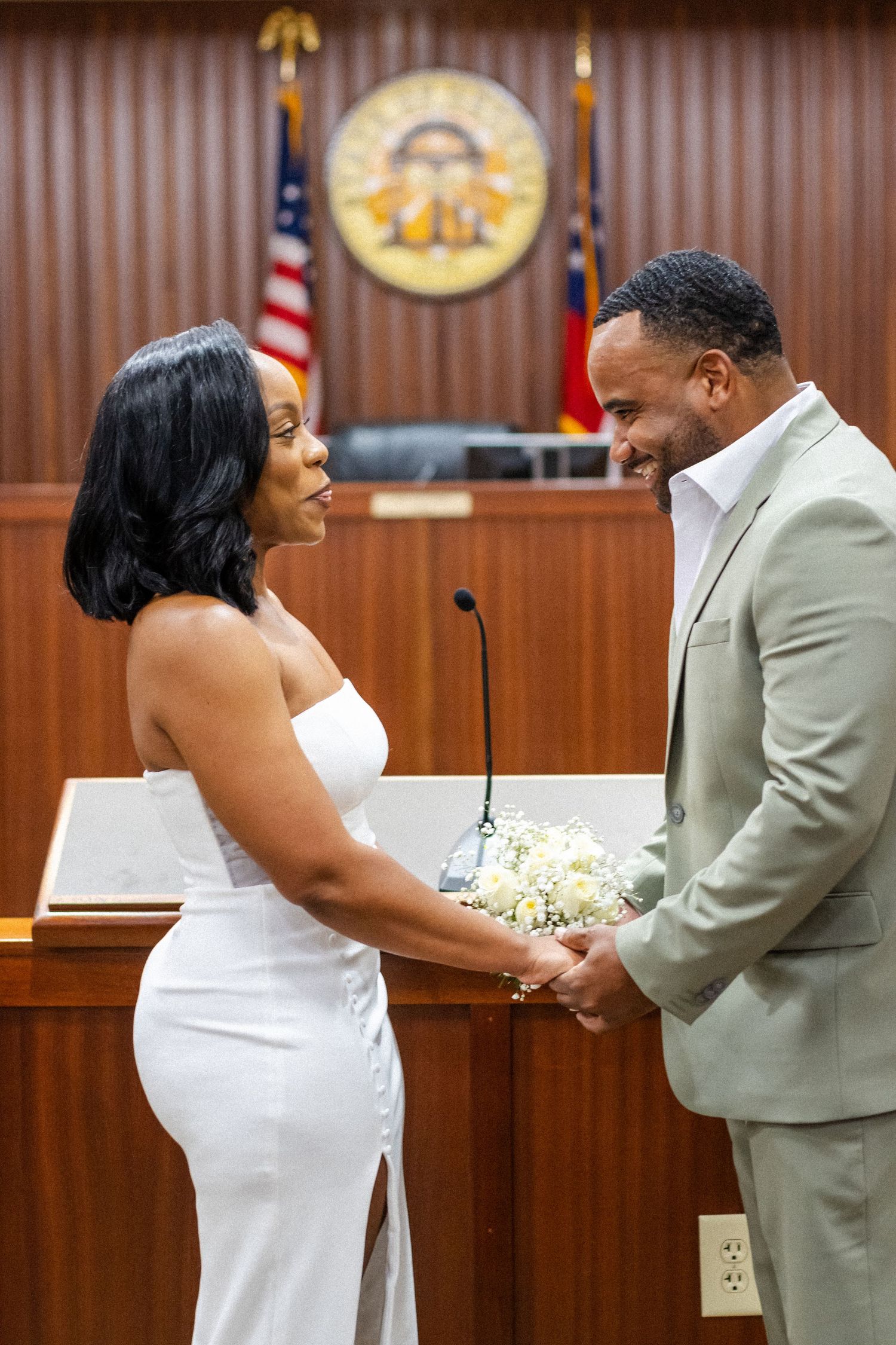A couple smiles at each other during their courthouse wedding ceremony, with a state seal and American flag visible behind them.