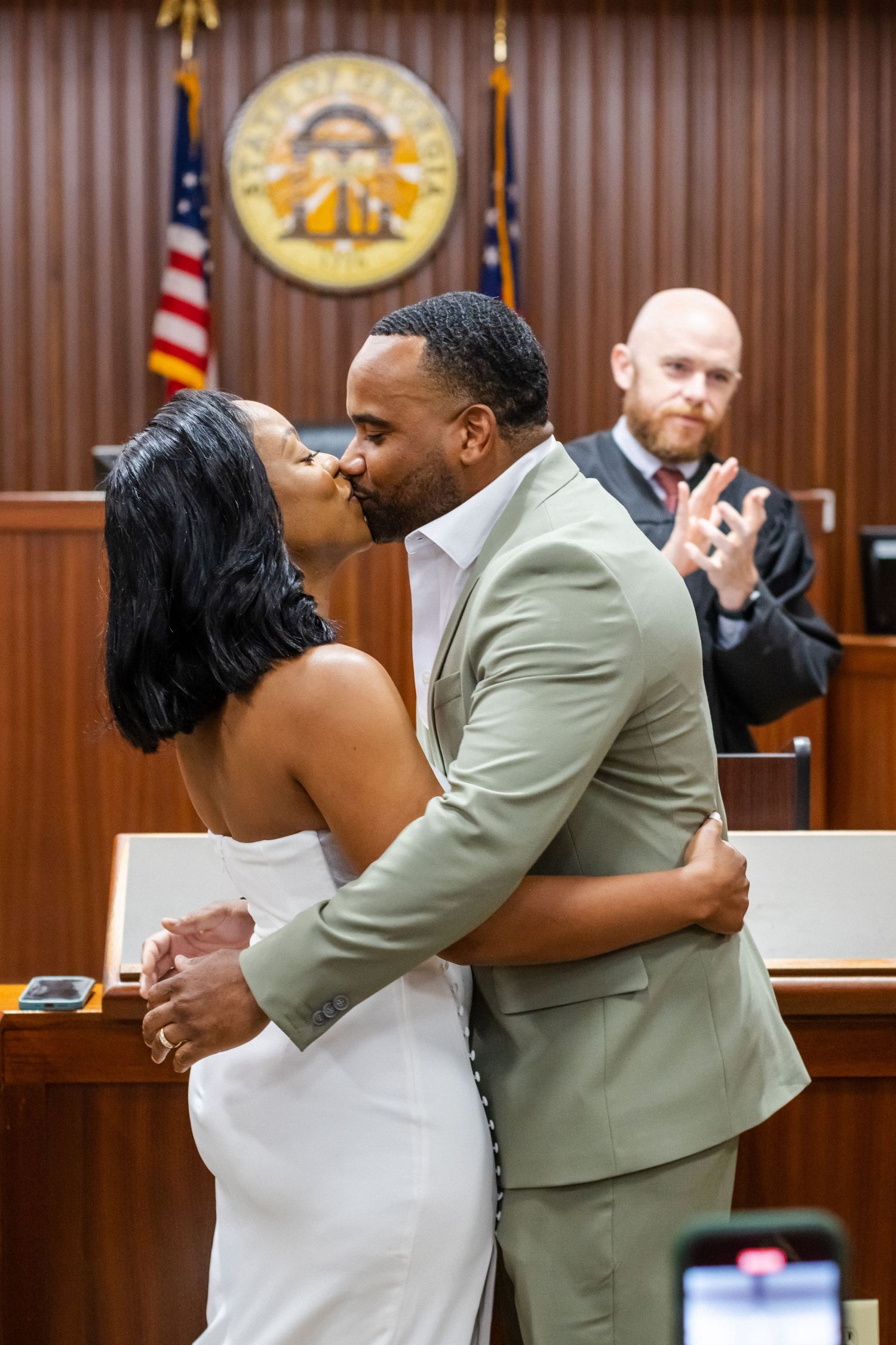 A couple shares their first kiss as newlyweds in a courthouse with an American flag and state seal visible in background.