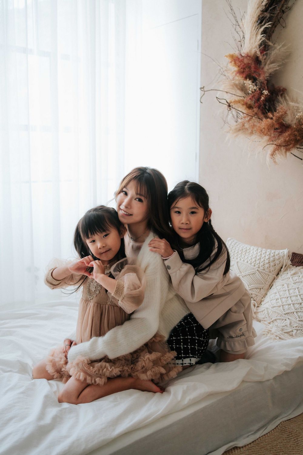 Friends relaxing together on a white bed wearing warm sweaters in a bright room.