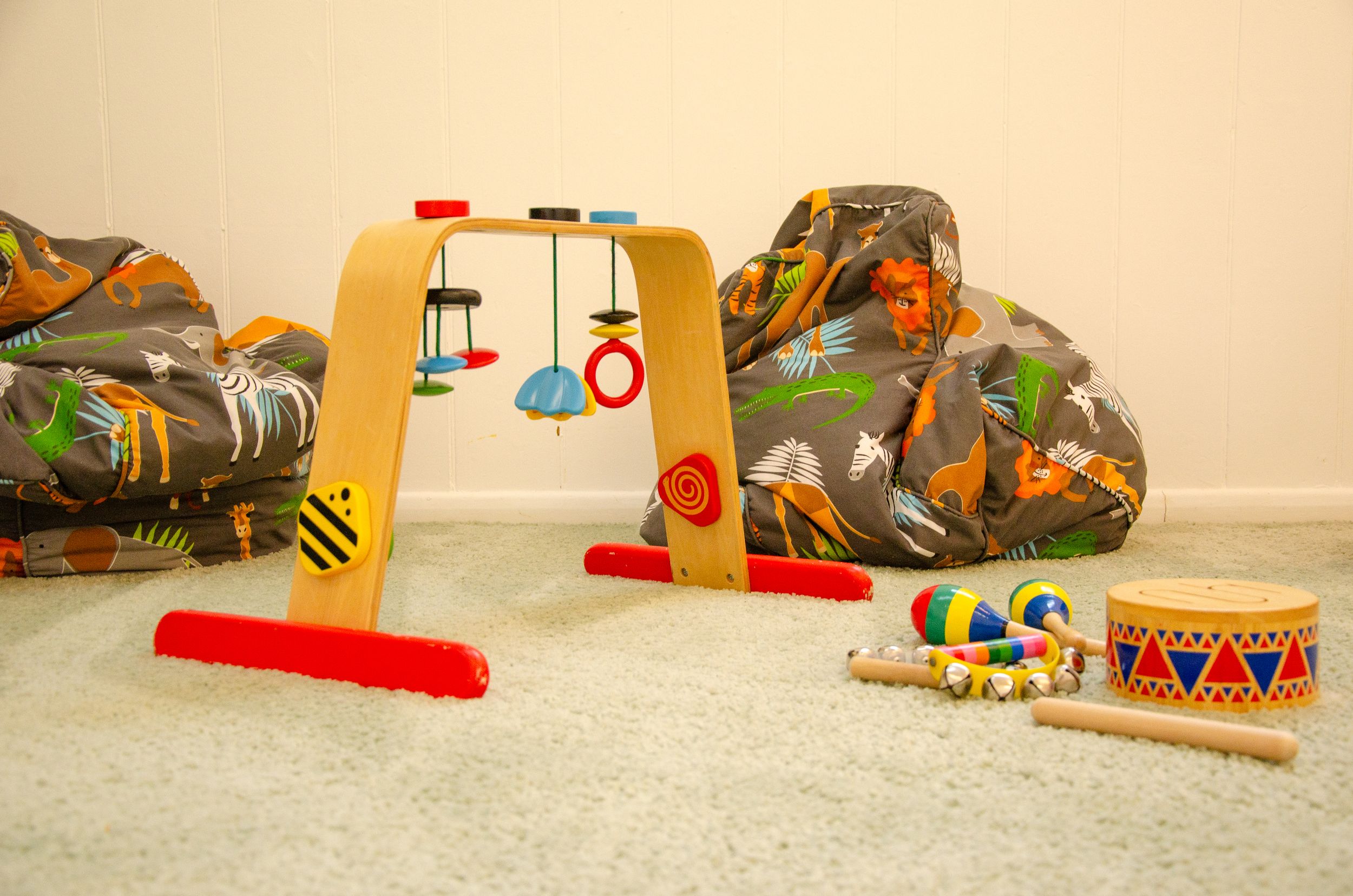 Colorful children's toys including a wooden balance beam, drum, and cushions arranged on carpet.