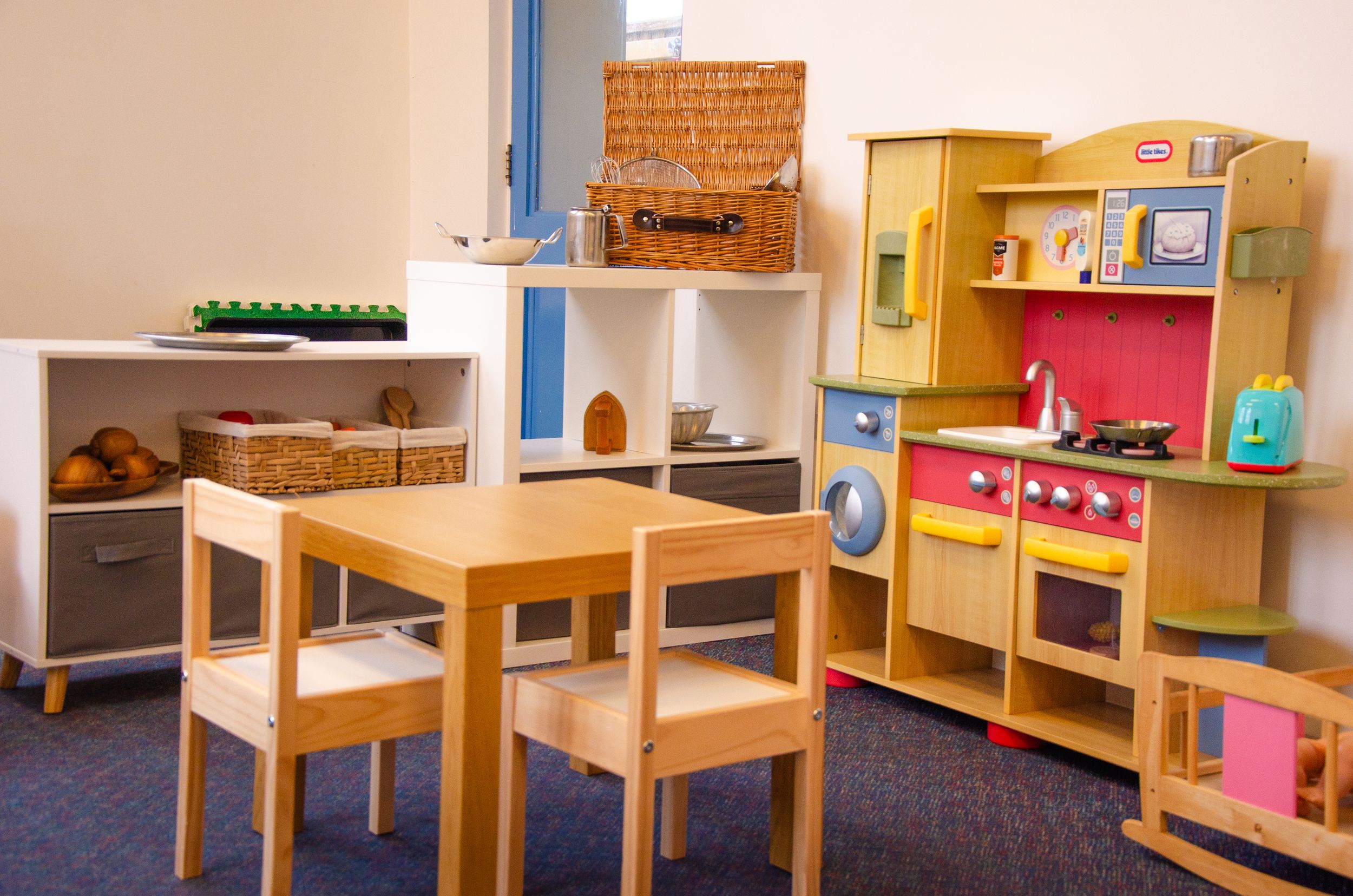 Educational playroom with wooden table, chairs and toy kitchen setup.