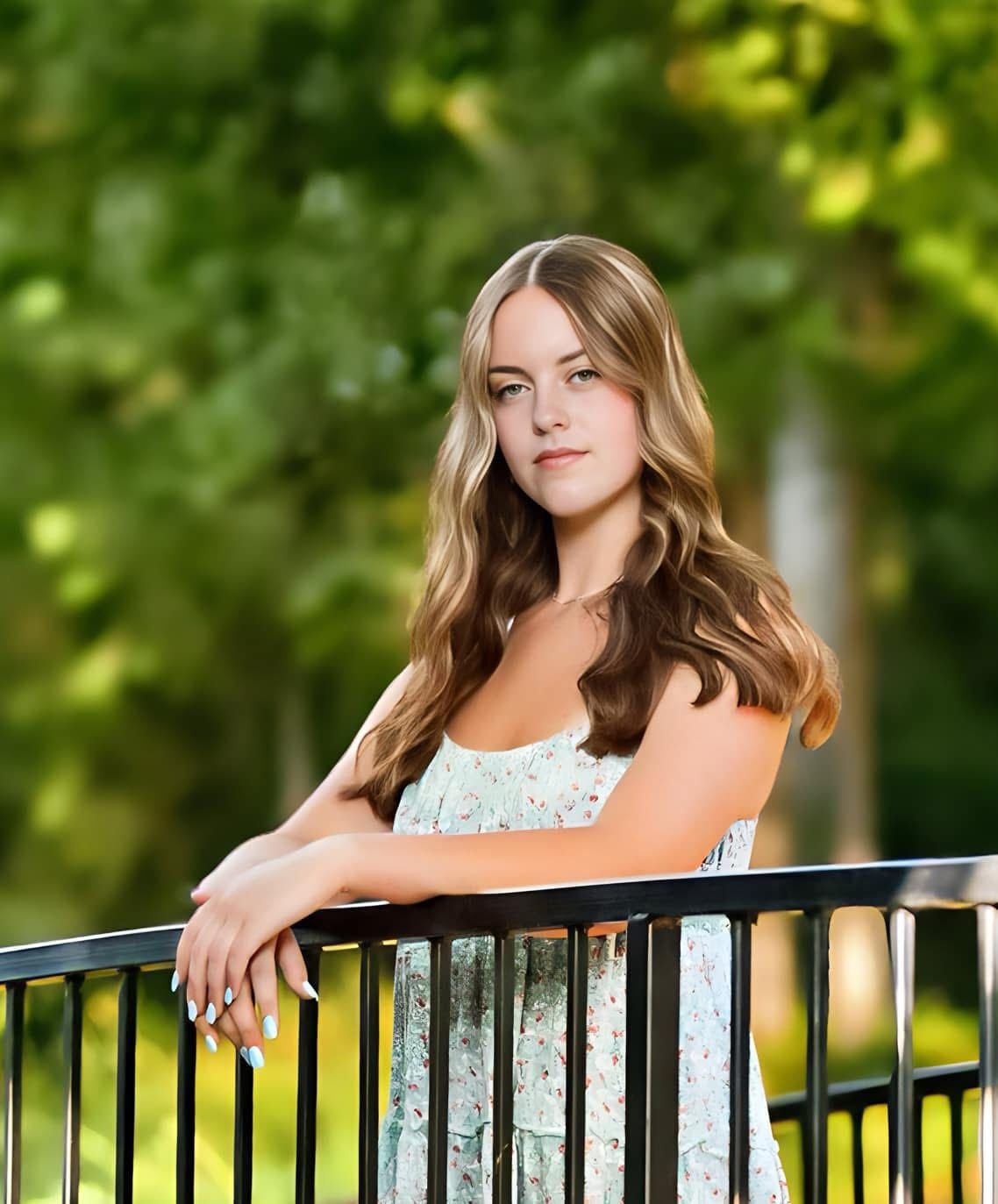 A person in a white lace dress poses against a black metal railing with sunlit trees in the background.