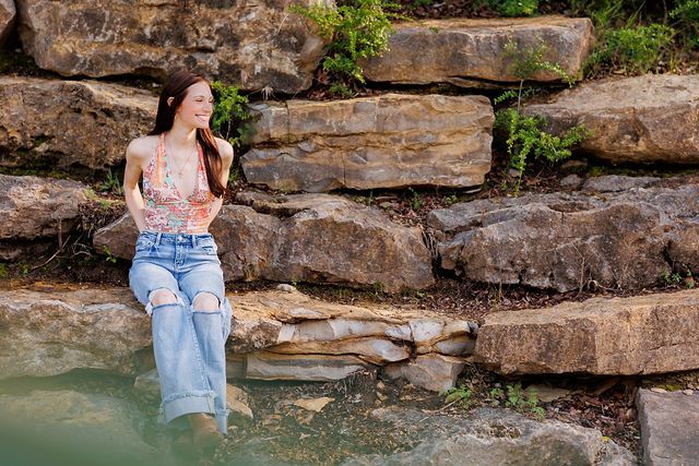 Artistic senior portrait by rocky waterfall wearing casual denim and pink top.