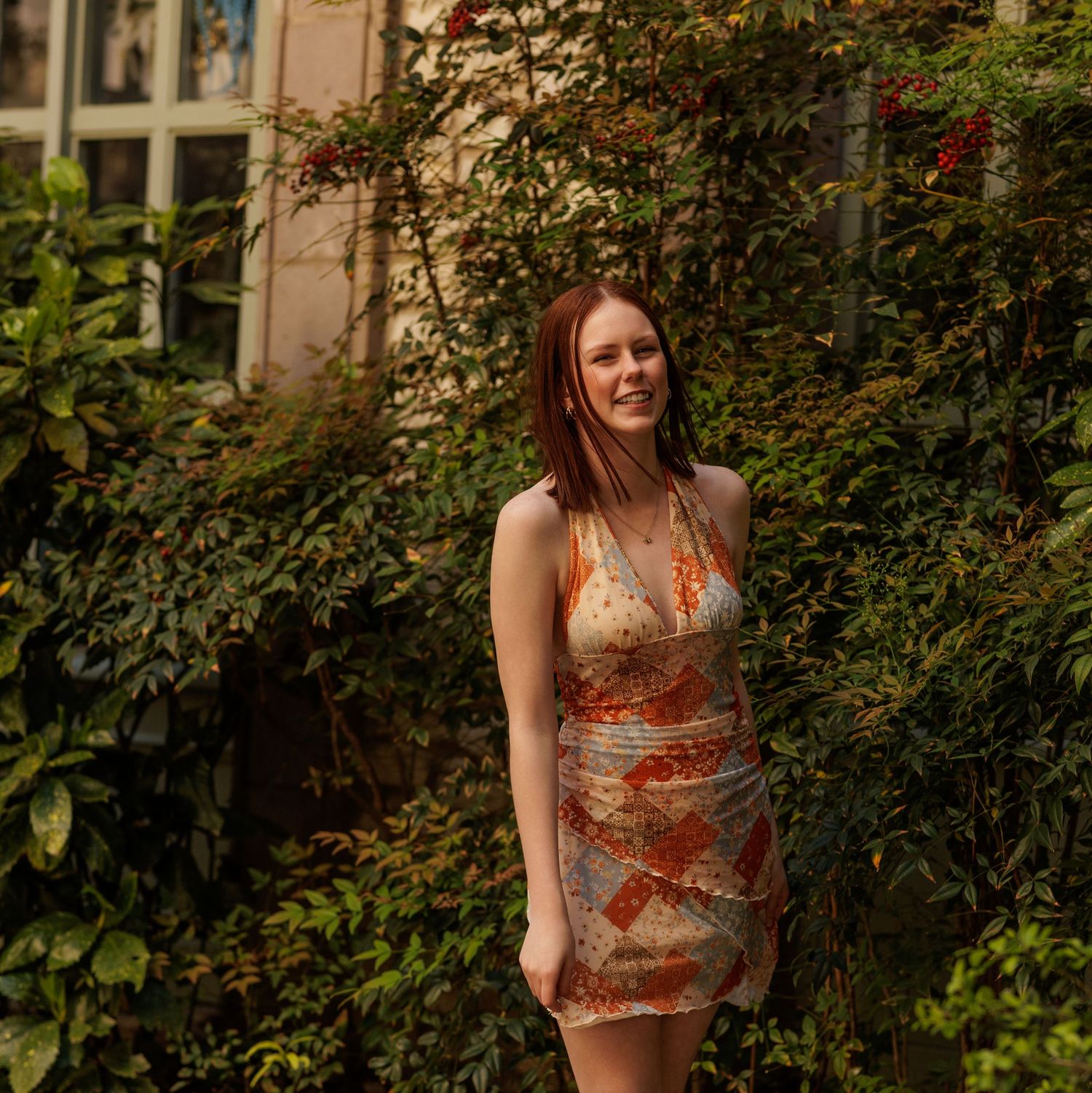 Young woman in floral mini dress smiling among lush green and red flowering vines outside a building.