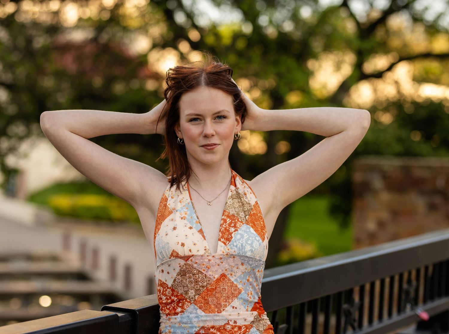 Young woman in floral halter dress poses outdoors near railing with hands in red hair, green trees behind her.