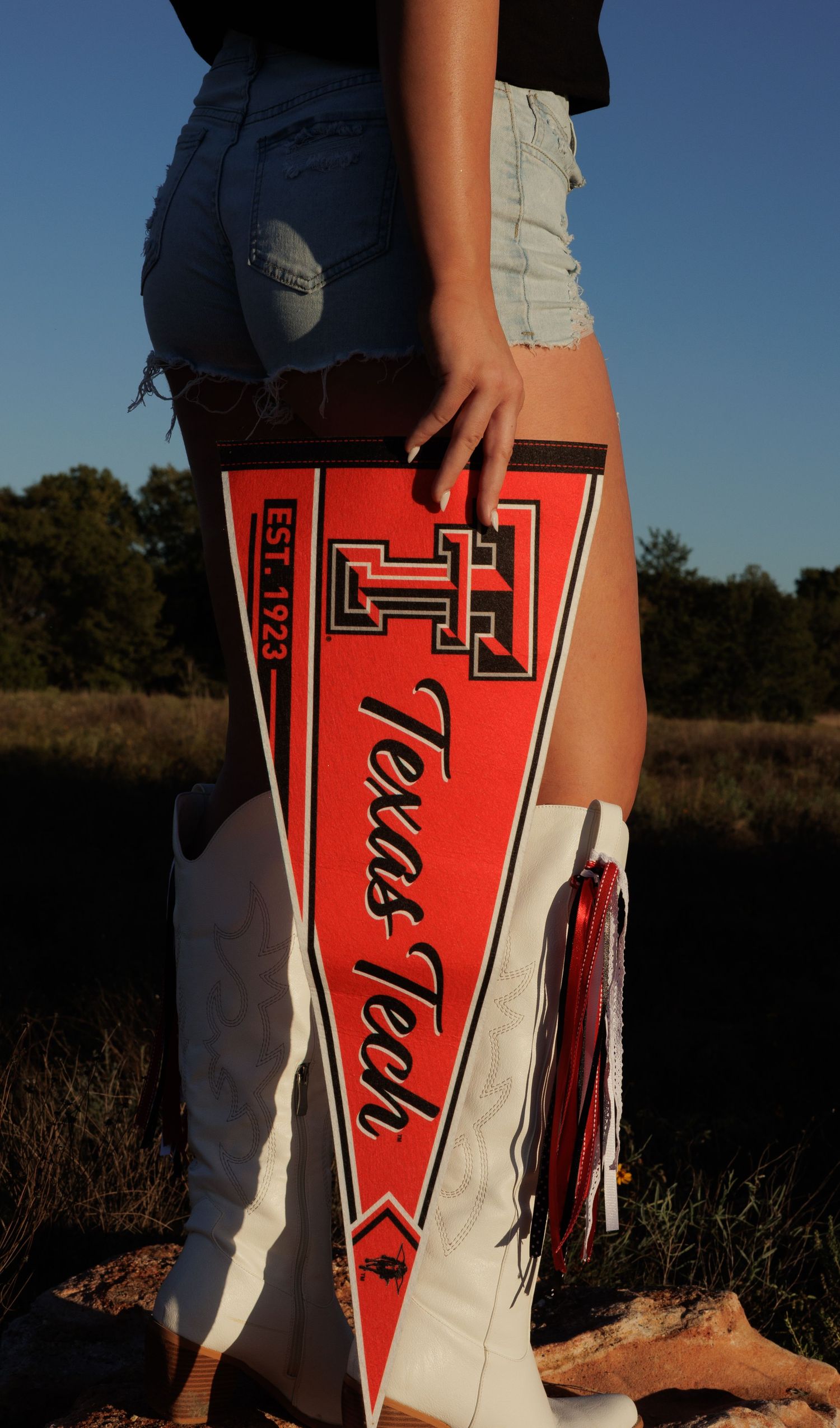 Woman in denim shorts and white boots holding a red Texas Tech University pennant flag outdoors.