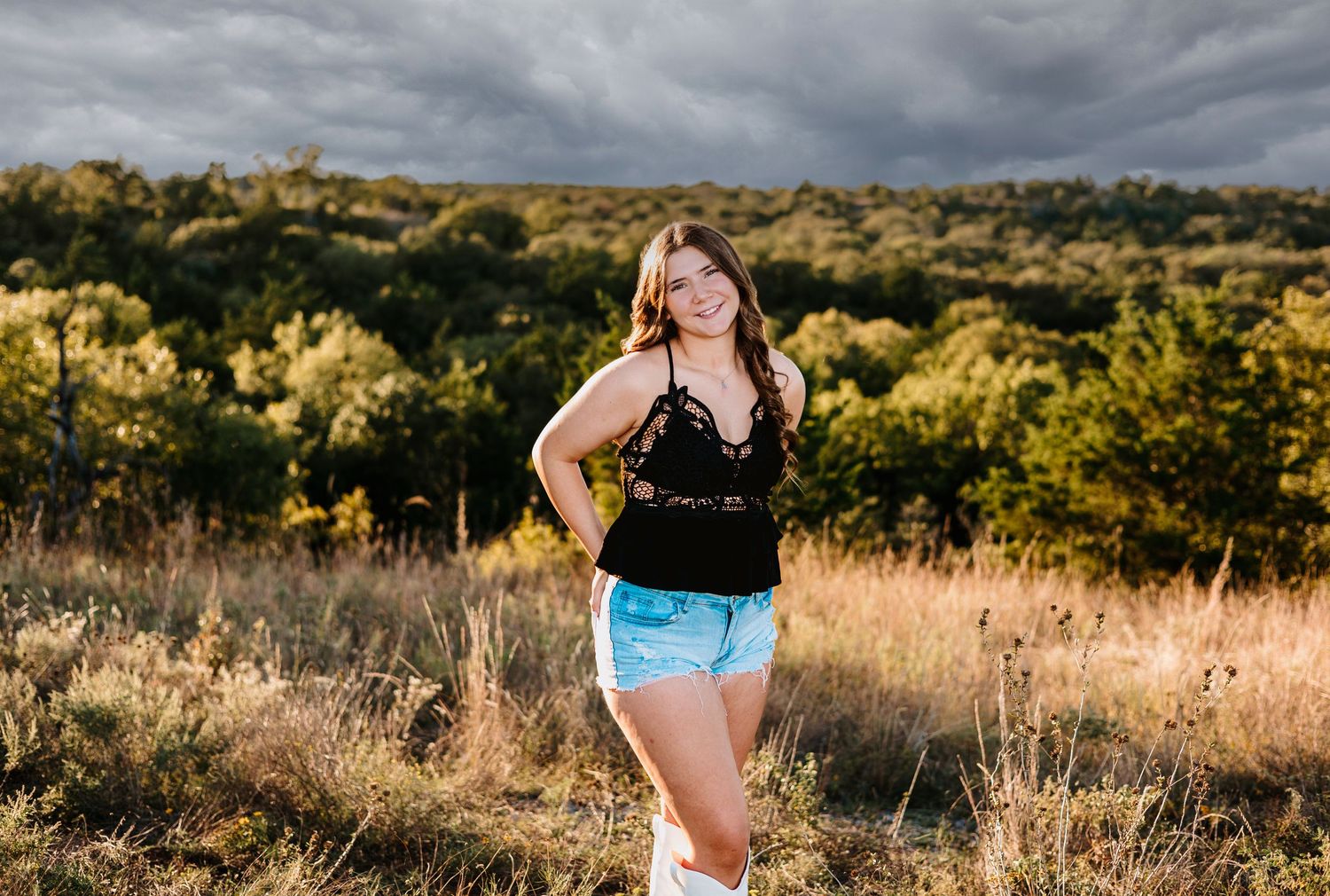 Young woman in black top and denim shorts posing in a golden field with dramatic stormy skies and trees.