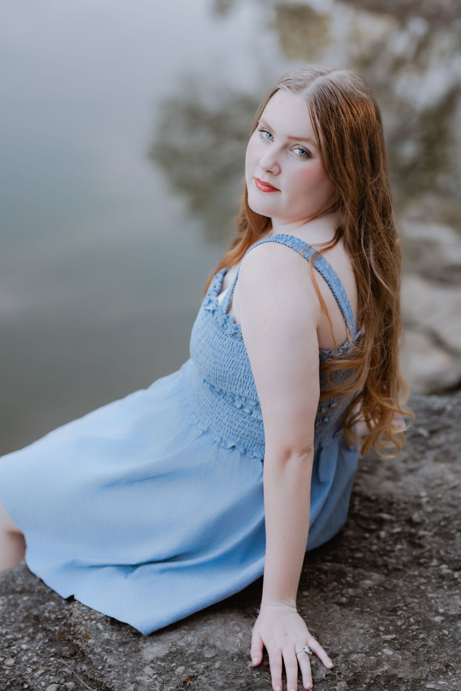 Young woman with long red hair in a light blue sundress sitting on rocks near a reflective body of water.