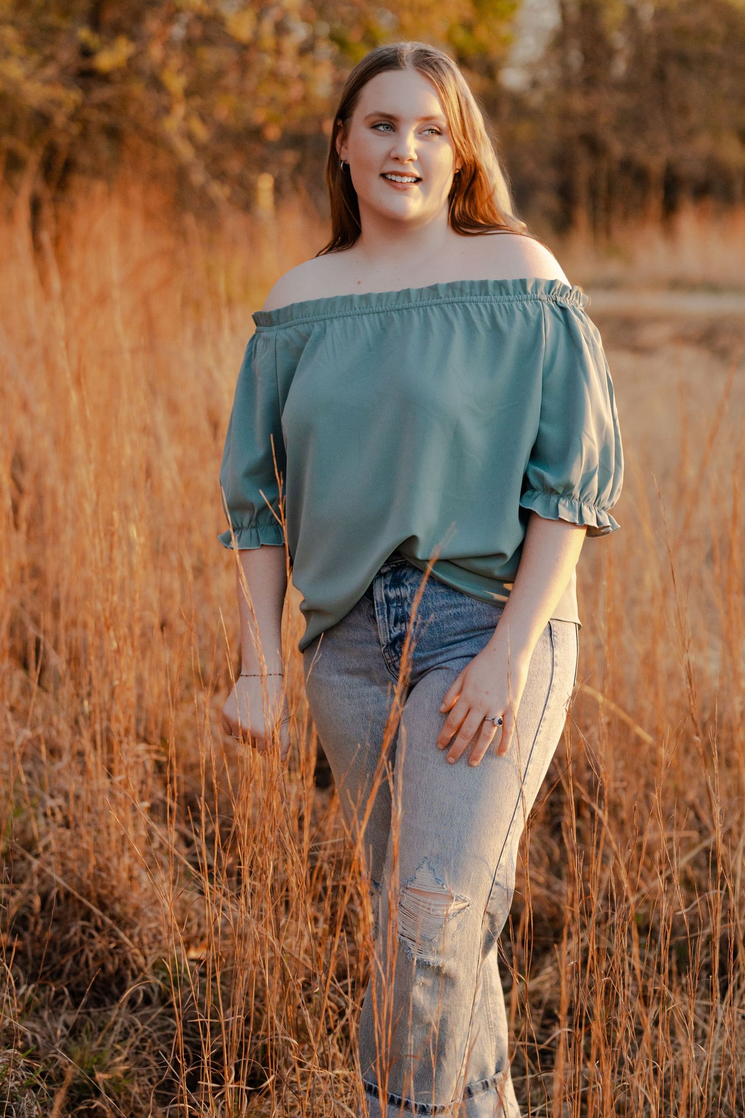Young woman in teal off-shoulder top and ripped jeans posing in golden field at sunset.