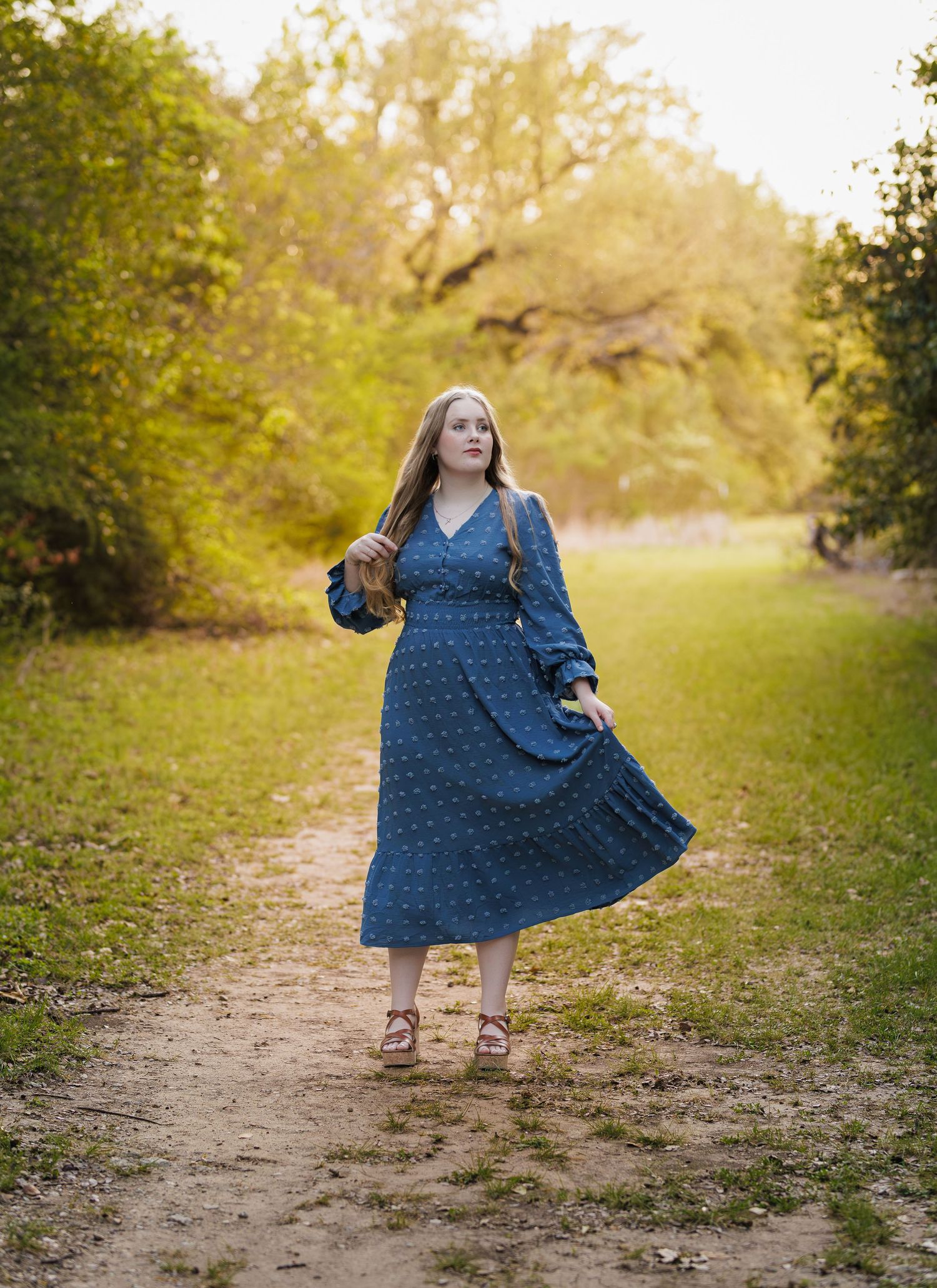 Young woman in blue polka dot midi dress walks a scenic dirt path surrounded by lush green trees in golden light.