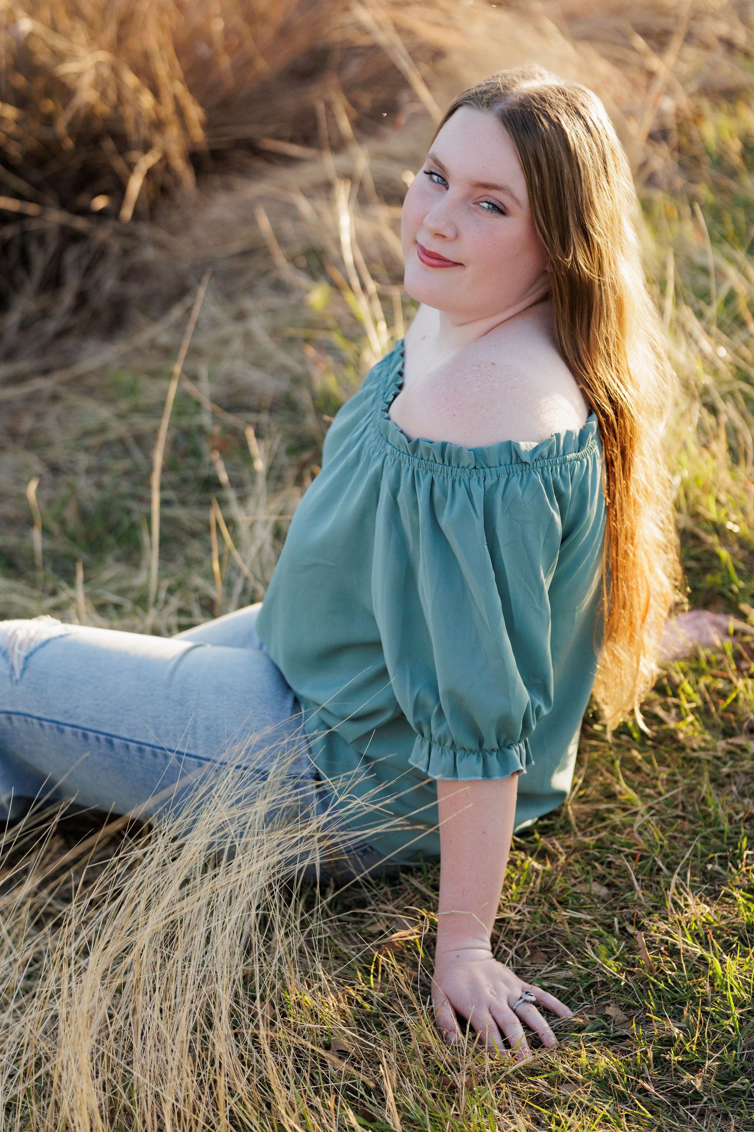 Young woman with red hair sits in golden grass field wearing teal off-shoulder top and jeans in warm sunset light.