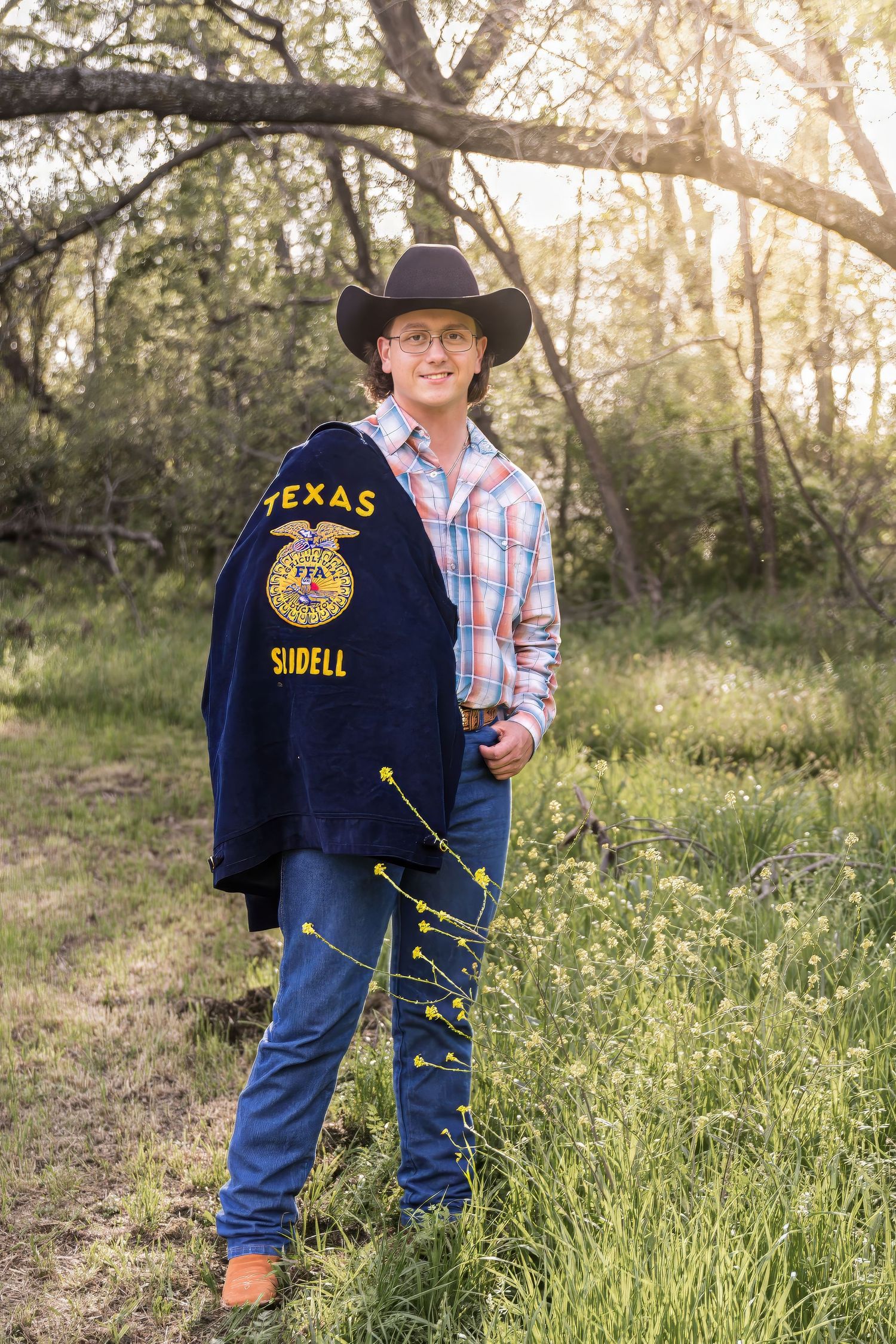 A seniorwearing a Texas FFA jacket and cowboy boots stands in a grassy field at sunset.