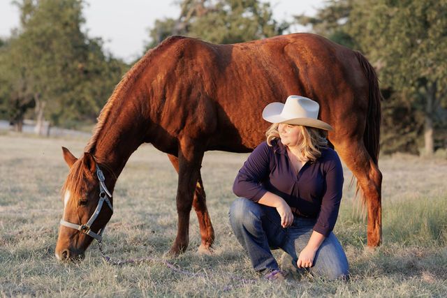 A brown horse grazes while someone in a cowboy hat sits nearby in a grassy field.